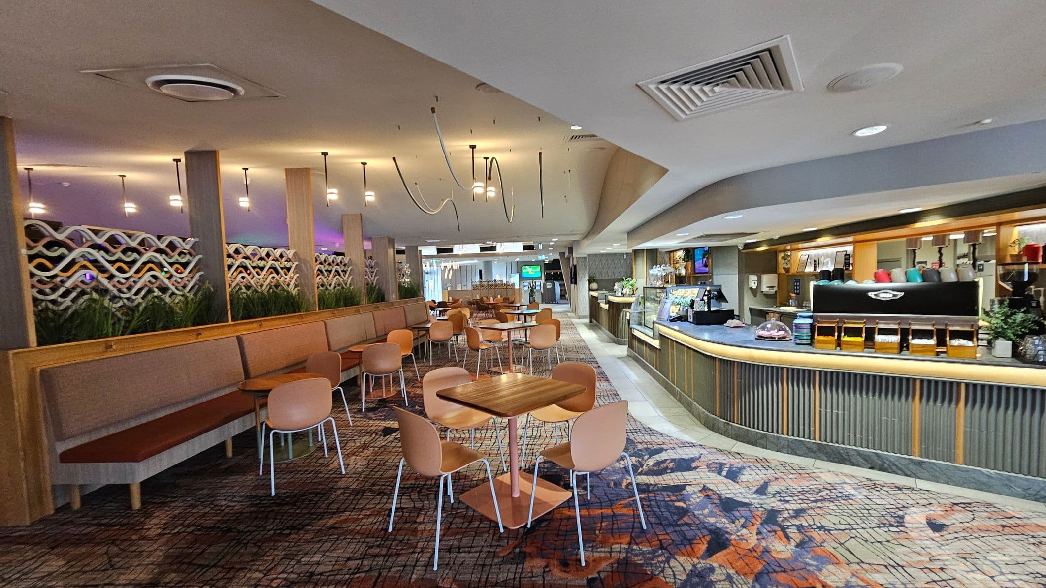 A modern café interior with a patterned carpet floor. On the left, there are long brown benches with small wooden tables and light brown chairs. Decorative wavy panels with plants are behind the seating. In the middle, more tables and chairs are arranged. On the right, a counter with a coffee machine, cups, and a display case. The ceiling has recessed lighting and hanging fixtures.