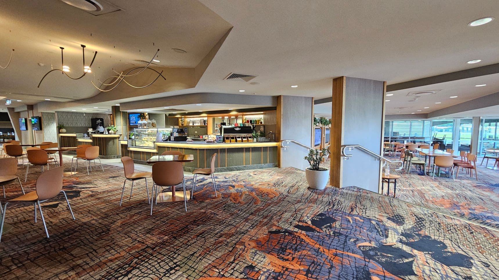 A spacious café interior with a modern design. On the left, there are several wooden tables and chairs on a patterned carpet with orange and black tones. In the centre, a counter with a coffee machine and display case is visible, surrounded by light wood panelling. On the right, more seating is arranged near large windows, allowing natural light to enter. The ceiling features abstract light fixtures, and a potted plant is placed near the railing.