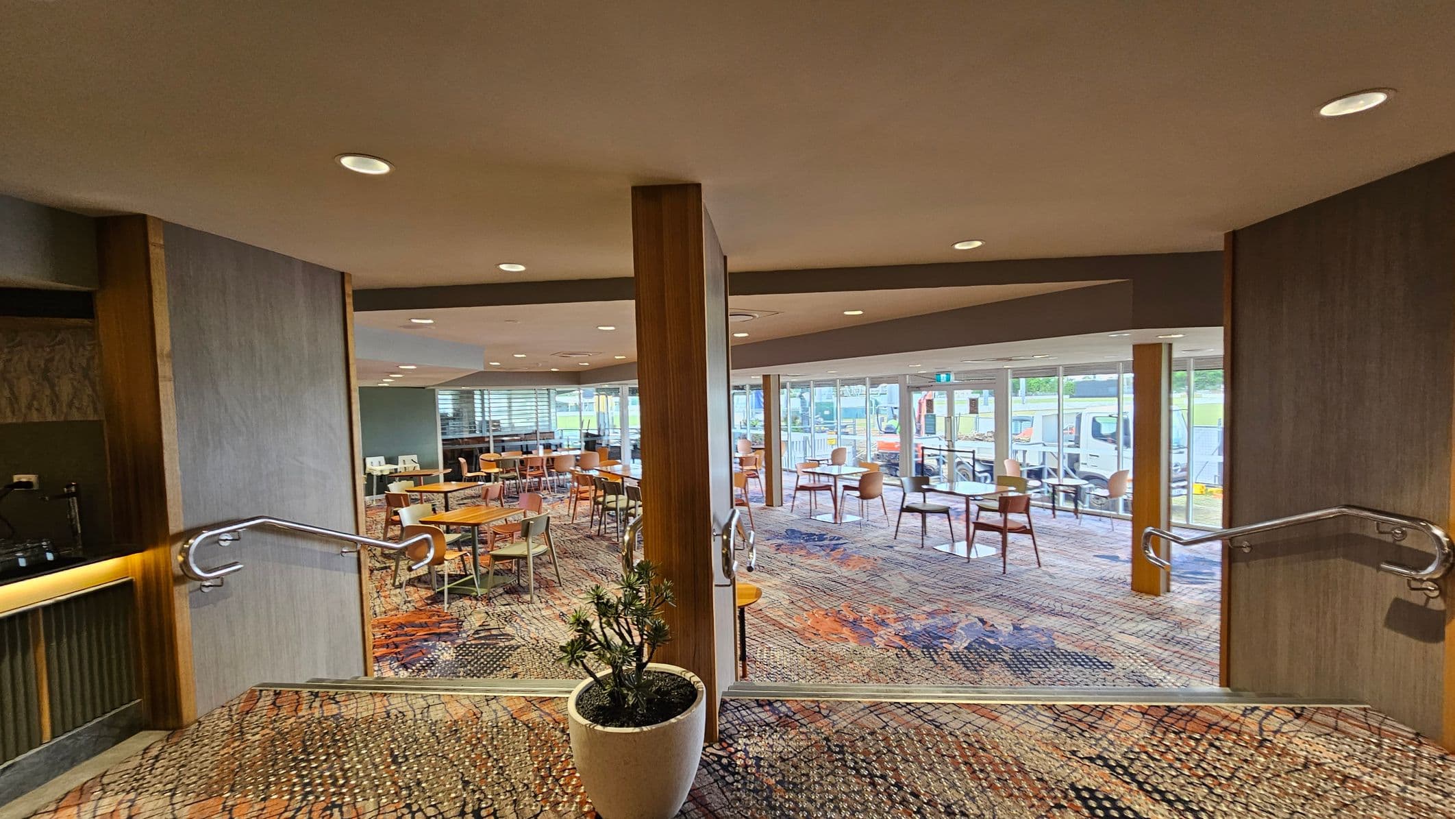 A spacious indoor area with patterned carpet flooring. In the foreground, a potted plant sits between two wooden pillars. The room features several wooden tables and chairs arranged throughout. Large windows in the background allow natural light to enter, revealing a view of parked vehicles outside. The ceiling is fitted with recessed lighting, and there are metal handrails on either side of the entrance.