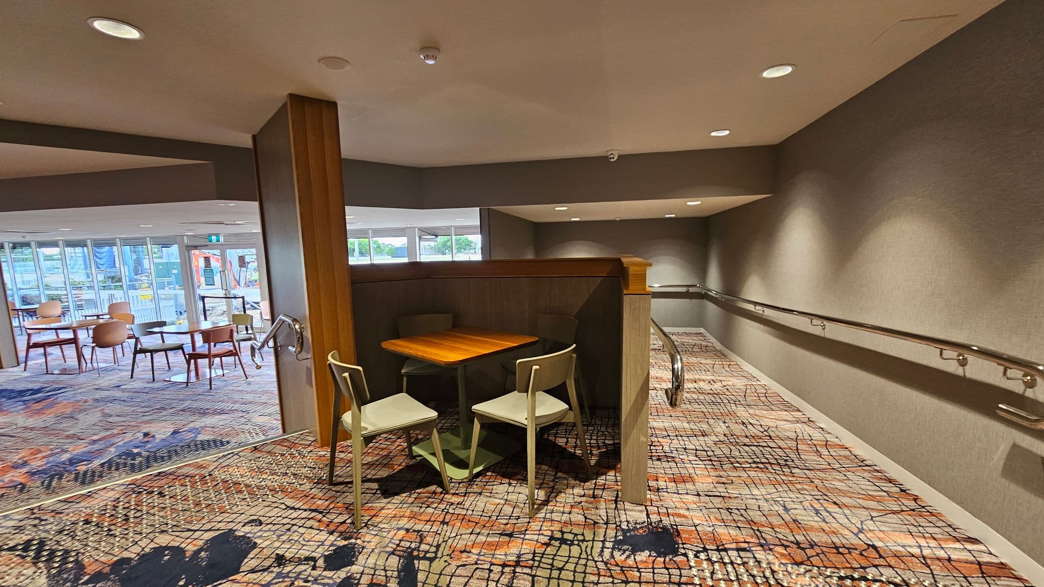 A modern interior space with a patterned carpet floor. On the left, there is a small wooden table with two chairs. Beyond that, a seating area with several tables and chairs is visible through large glass windows. On the right, a ramp with metal handrails leads down a corridor. The ceiling is fitted with recessed lighting, and the walls are painted in a neutral tone.