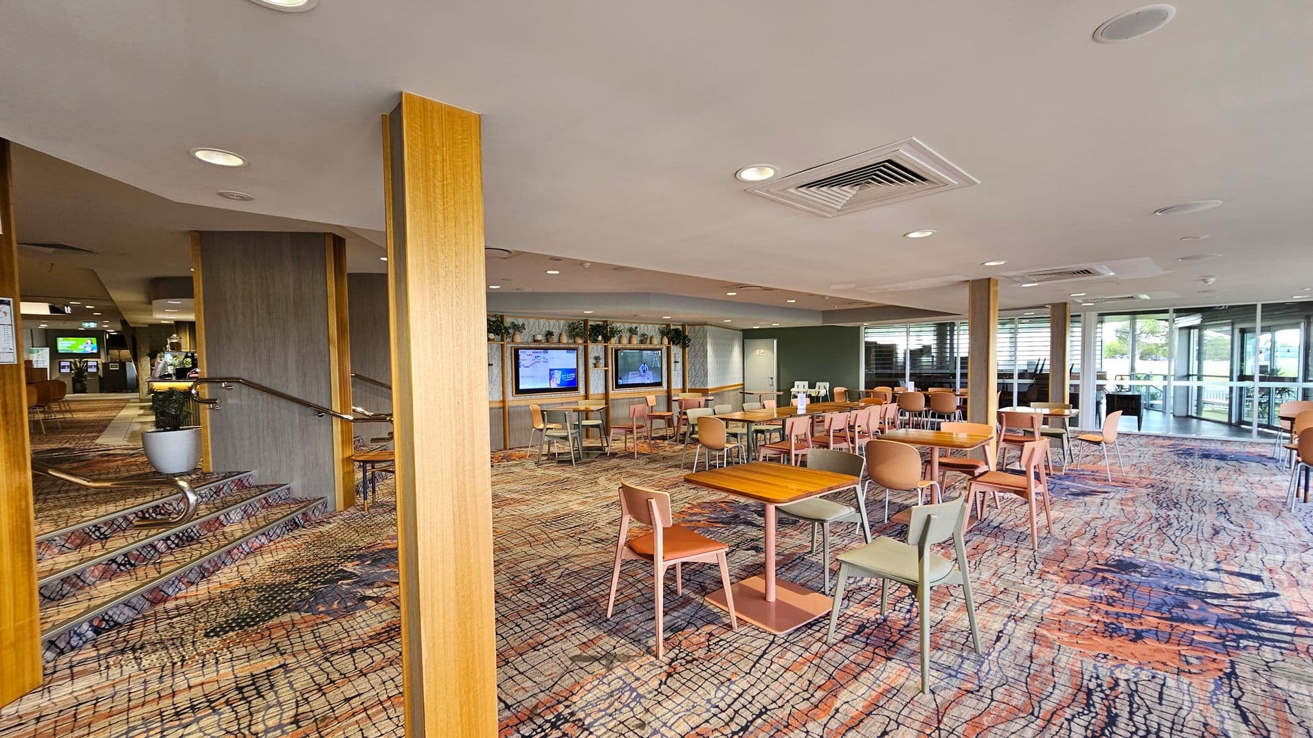 A spacious interior with a patterned carpet floor featuring abstract designs in orange, blue, and grey. On the left, there are steps with a metal railing and a potted plant. The middle area has wooden tables and chairs arranged in rows. The back wall displays two screens with greenery above them. On the right, large windows with blinds allow natural light in. The ceiling is fitted with recessed lighting and air vents.