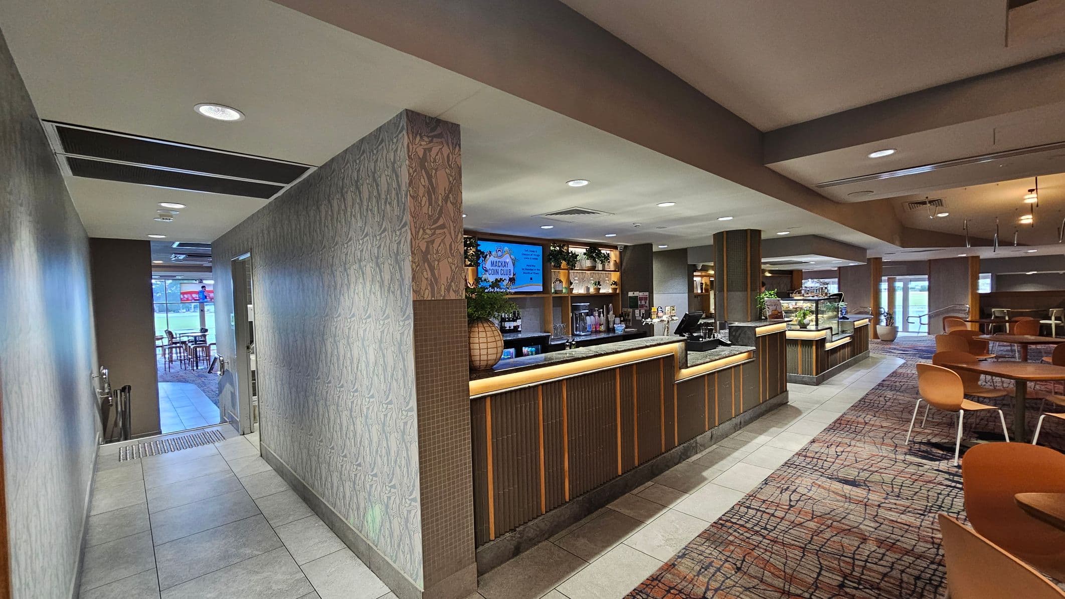 A modern interior with a bar area in the centre, featuring a wooden counter and a digital screen above displaying information. The left side has a hallway with patterned wallpaper and tiled flooring leading to a seating area with visible tables and chairs. On the right, there are round tables with orange chairs on a patterned carpet. The ceiling is fitted with recessed lighting, and large windows in the background allow natural light to enter.