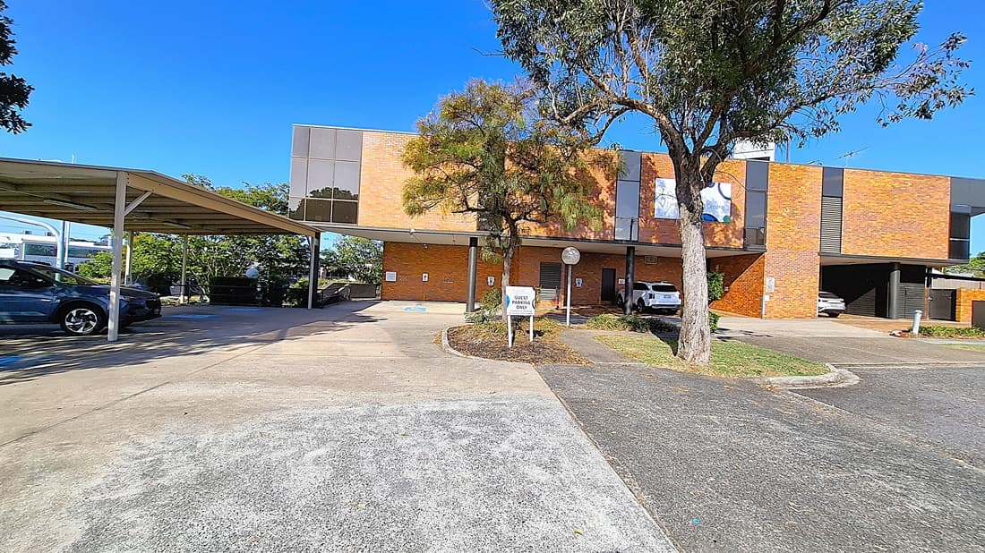 A car park with a covered area on the left, housing several parked cars. In the middle, a tree stands in front of a two-storey brick building with large windows. The building has a sign and a circular driveway leading to the entrance. On the right, more parking spaces are visible, with additional cars parked. The ground is a mix of concrete and asphalt, and the area is surrounded by greenery.