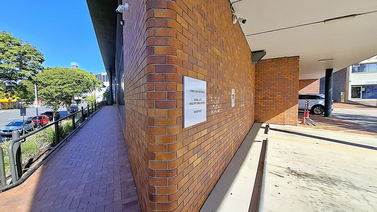 On the left, a brick walkway with a black metal railing runs alongside a row of trees and parked cars. In the middle, a brick wall with signs and a security camera. On the right, a covered parking area with a concrete floor, a black column, and a parked car. The ceiling is white with light fixtures and seams.