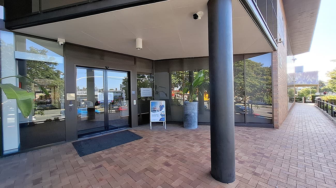 On the left, large glass windows reflect the surrounding trees and buildings. In the middle, a glass entrance door with a black frame, flanked by a signboard and a large potted plant. On the right, a black column supports the overhang, with more glass windows and a brick walkway extending along the building. The floor is made of red bricks, and the ceiling is white with light fixtures and a security camera.