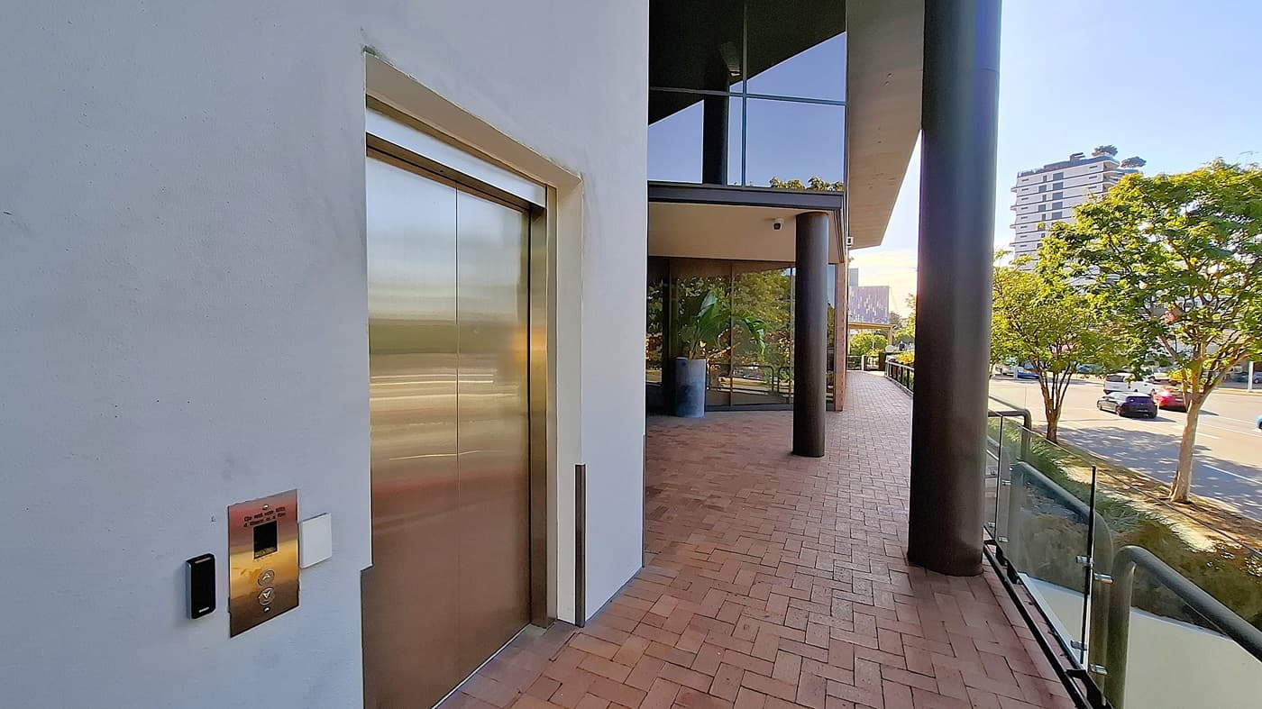 On the left, a stainless steel lift door set in a white wall with a control panel beside it. In the middle, a large black column supports an overhang, with glass windows and a potted plant visible. On the right, a brick walkway bordered by a glass railing, leading to a street with trees and parked cars. The floor is red brick, and the ceiling is white with recessed lighting.