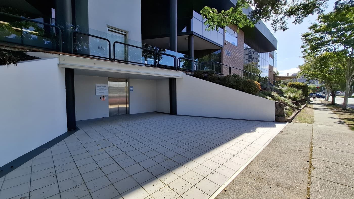 On the left, a white wall with a black column and a glass railing above. In the middle, a stainless steel lift door with a control panel and signs on either side. On the right, a sloped garden area with shrubs and a black railing, leading to a brick building with large windows. The floor is tiled with light-coloured square tiles, and the ceiling above the lift area is white.