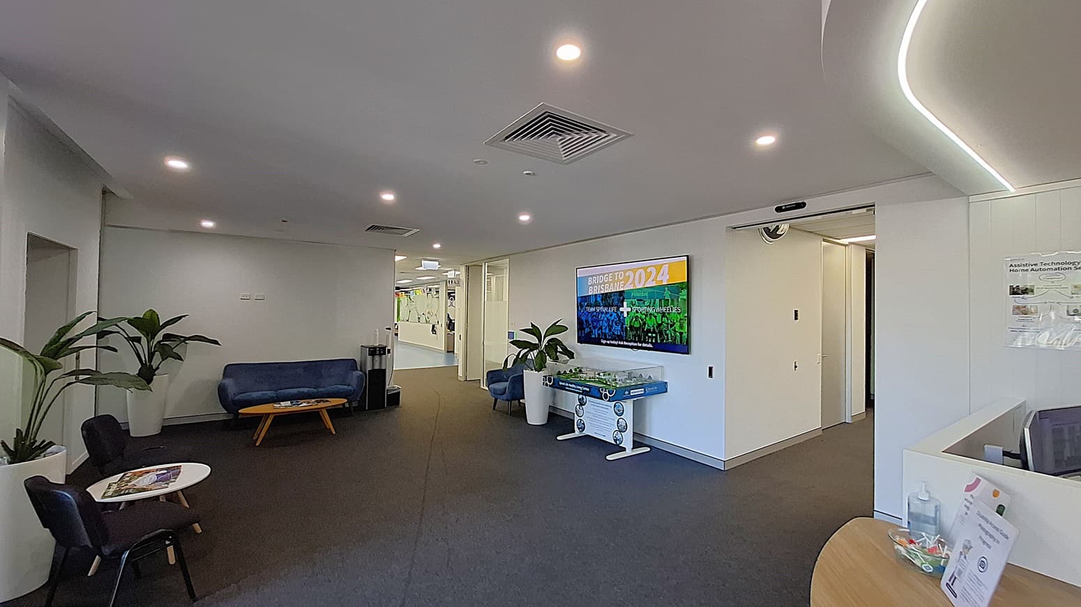 On the left, two black chairs and a small round table with magazines, next to a large potted plant. In the middle, a blue sofa with a wooden coffee table in front, and a hallway leading to another area. On the right, a wall-mounted TV displaying "Bridge to Brisbane 2024," a foosball table, and another potted plant. The floor is covered with dark carpet, and the ceiling is white with recessed lighting and air vents.