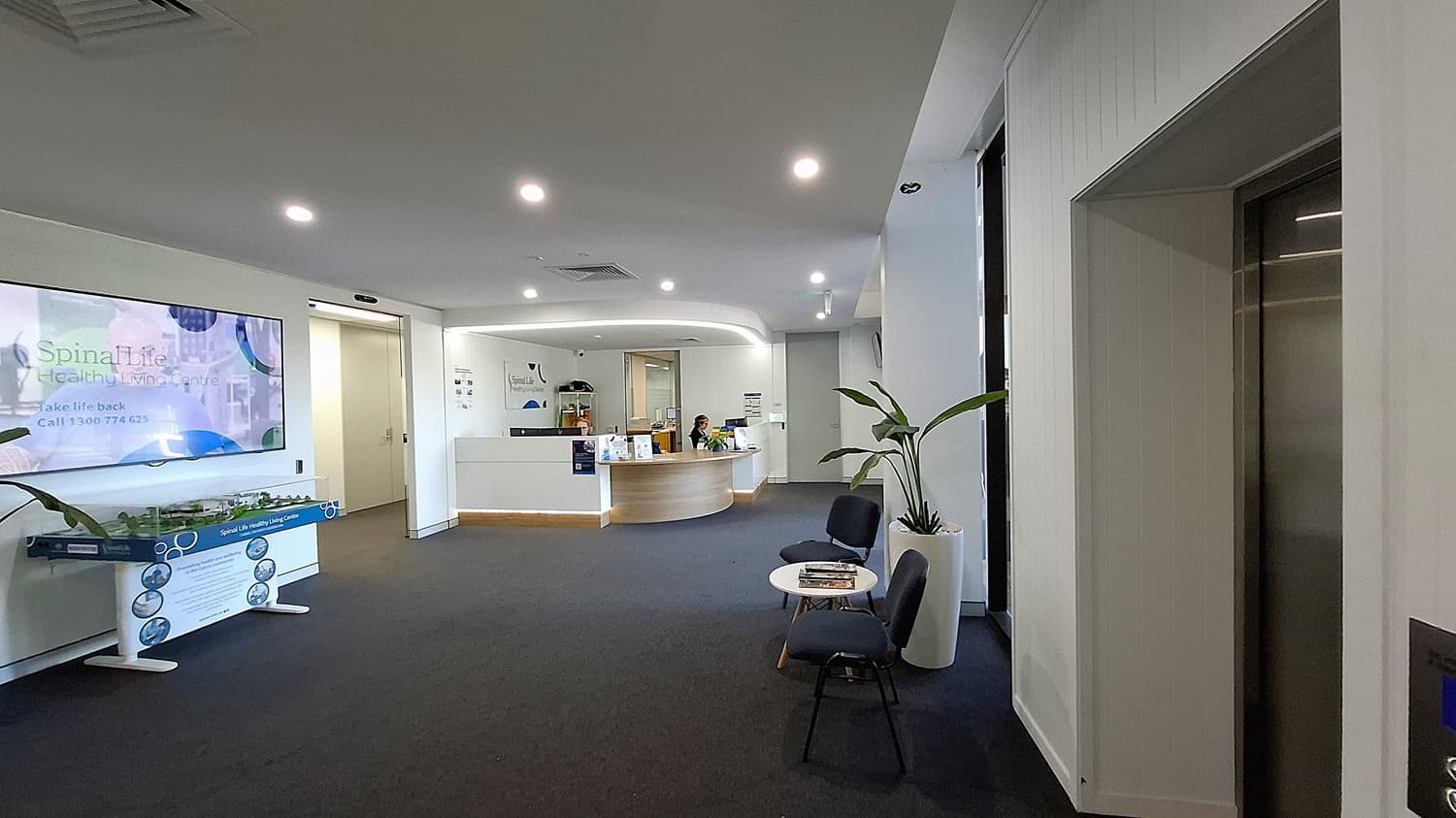 On the left, a large wall-mounted TV displaying "Spinal Life Healthy Living Centre" above a model display table. In the middle, a curved reception desk with two people seated behind it, computers, and informational brochures. On the right, a lift with stainless steel doors, a potted plant, and two black chairs with a small round table holding magazines. The floor is covered with dark carpet, and the ceiling is white with recessed lighting and air vents.