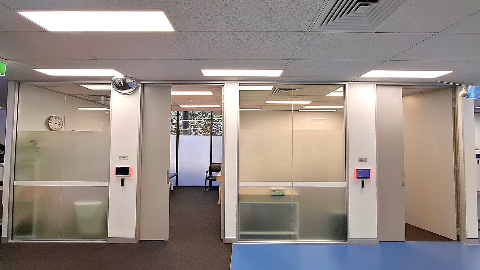On the left, a frosted glass door with a wall-mounted clock inside the room. In the middle, an open doorway leading to a room with a chair and a table, and a window with frosted lower panels. On the right, another frosted glass door with a blue-lit access panel and a partially visible hallway. The floor is a mix of dark carpet and blue vinyl, and the ceiling is white with recessed lighting and air vents.