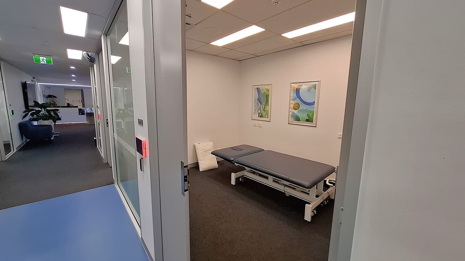 On the left, a hallway with a blue floor and glass walls, leading to a reception area with a blue sofa and potted plants. In the middle, an open door reveals a room with a grey examination table, two framed abstract artworks on the wall, and a white pillow on the floor. On the right, a white wall with part of the door frame visible. The floor in the room is covered with dark carpet, and the ceiling has white panels with recessed lighting.