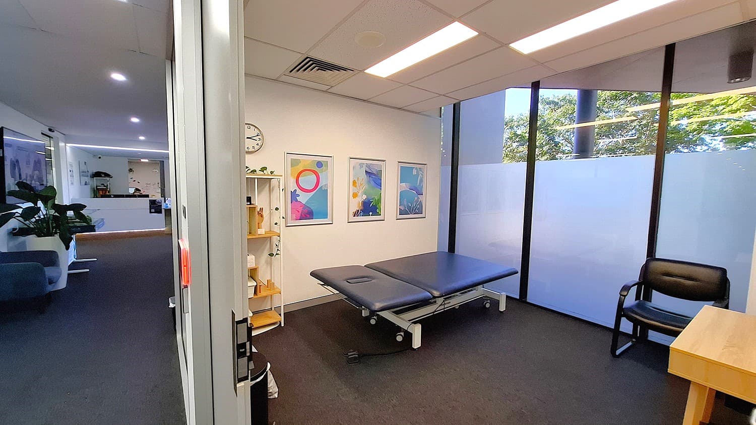 On the left, a hallway with dark carpet, a blue sofa, and potted plants. In the middle, a room with a black examination table, three framed abstract artworks on the wall, a white clock, and a white shelving unit with various items. On the right, large windows with frosted lower panels, a black chair, and a wooden desk. The ceiling has white panels with recessed lighting and air vents.