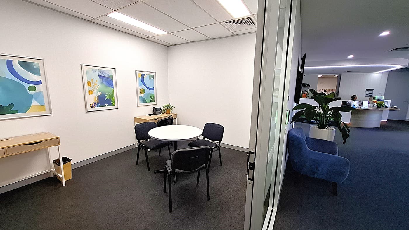 On the left, a small wooden table with a drawer and a bin underneath, three framed abstract artworks on the wall, and a round white table with four black chairs. In the middle, a glass door leading to a hallway. On the right, a blue sofa and a large potted plant. In the background, a curved reception desk with a person seated behind it and informational brochures. The floor is covered with dark carpet, and the ceiling has white panels with recessed lighting and air vents.