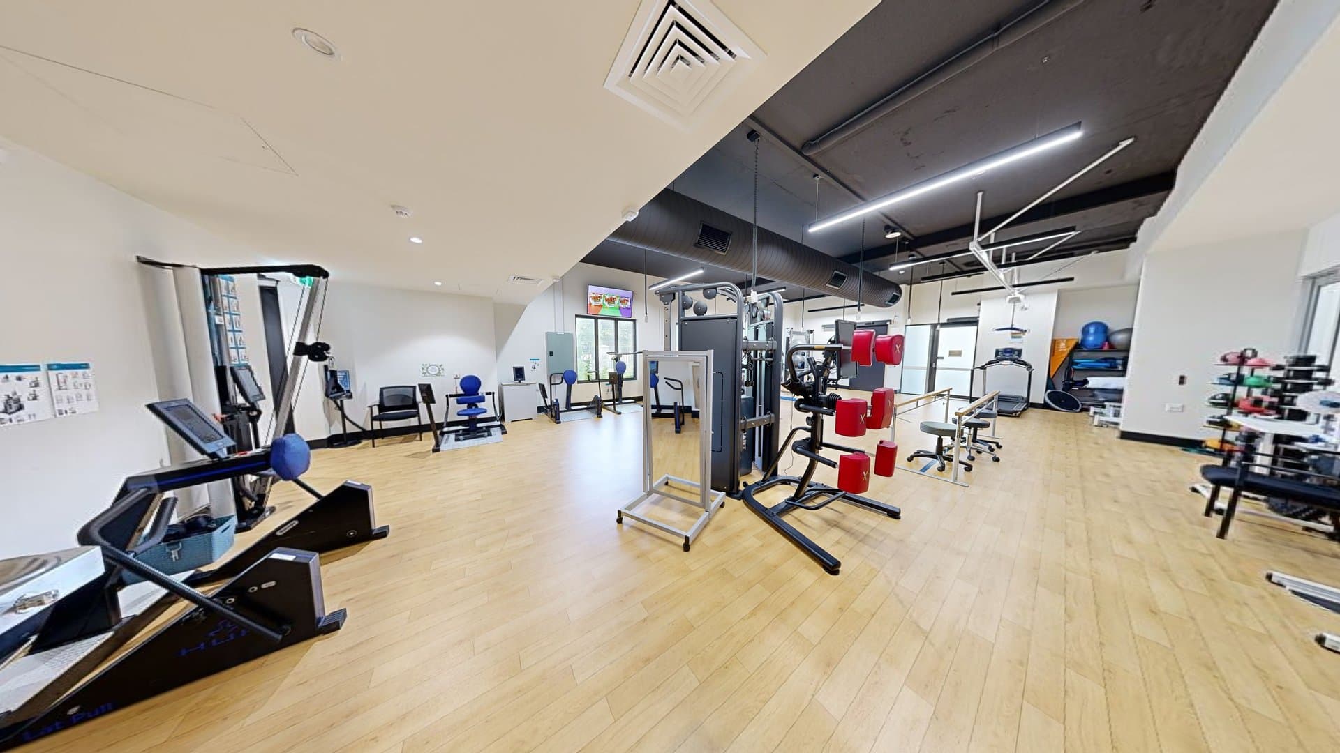 A lobby leading to the gym. Green doors on the right lead to Suites 3 and 4. On the left, an outward-opening glass door with affixed posters. A hand sanitiser machine is placed before it. Inside the gym, machines are arranged. The floor is timber, and the ceiling and walls are all white.