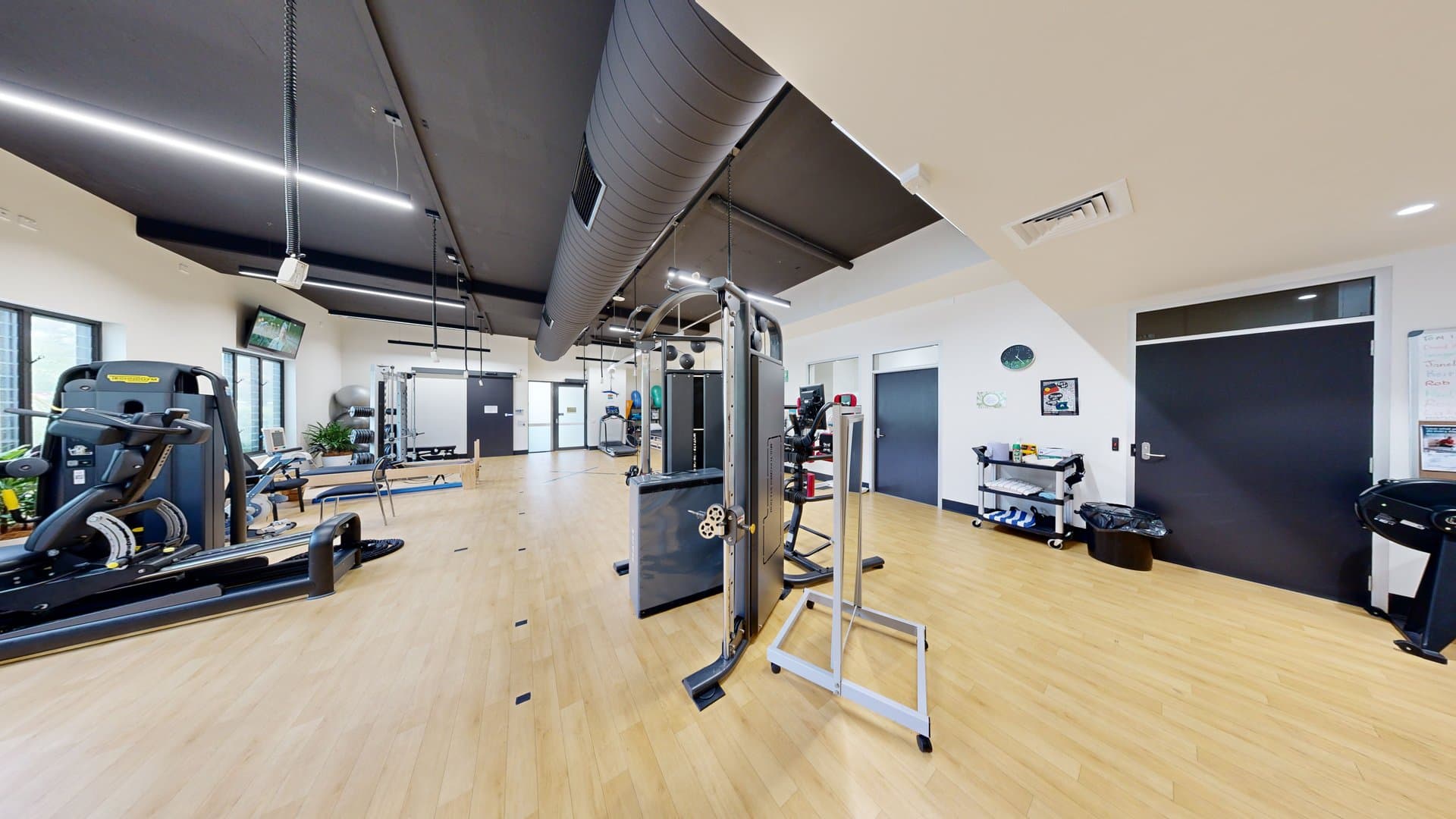 Inside the gym, a treadmill machine and bench machines are on the left, with a lat pull-down machine in the middle. The floor is timber, the walls are white with posters, and blue doors are on the right. A cart holding towels and cleaning materials sits beside a black rubbish bin against the right wall. A wall clock and poster adorn the wall above. The ceiling is black with strips of fluorescent lights and large aircon vents.