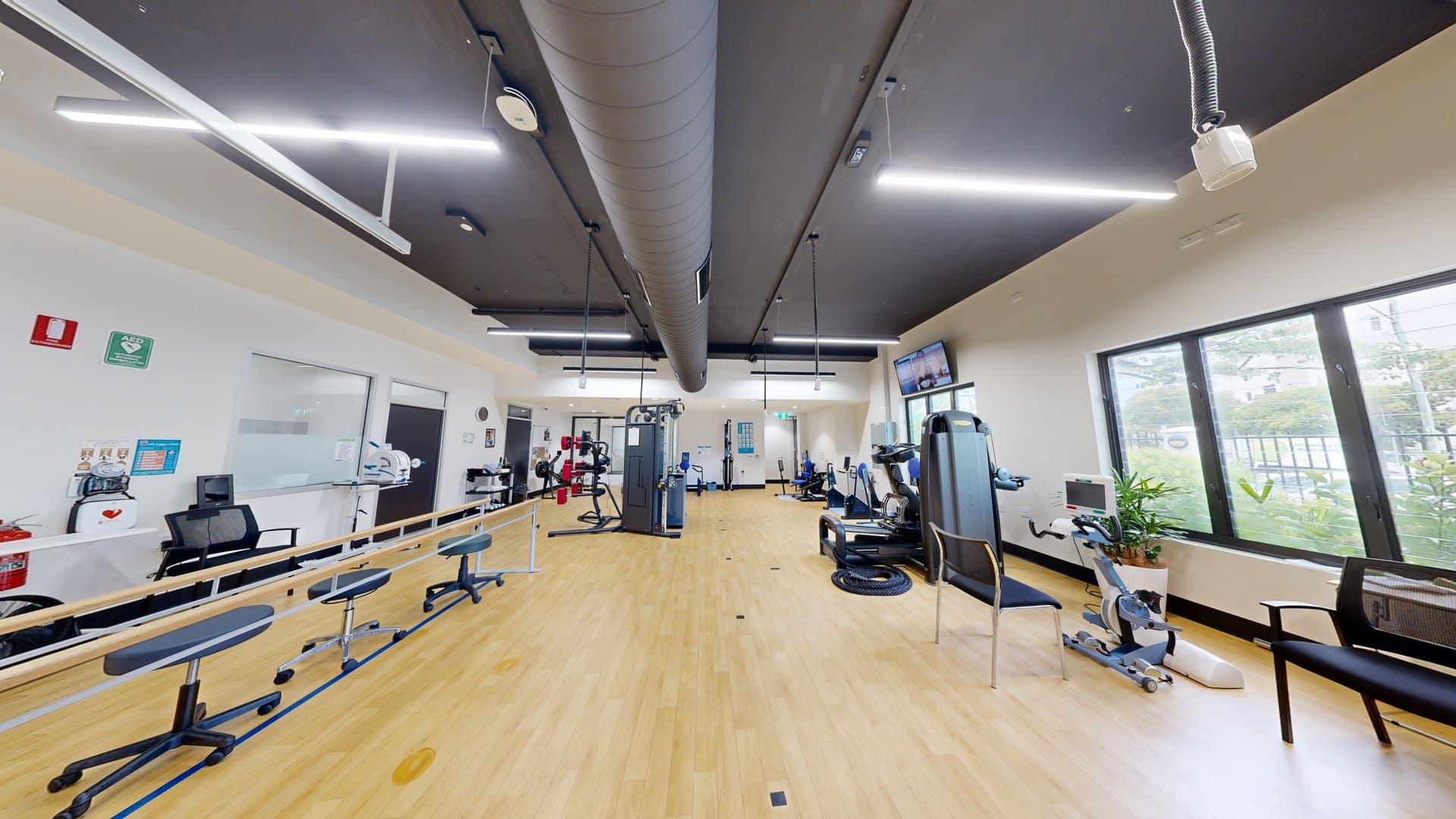 To the right is a bench chair positioned against a glass window, with Physiotherapy equipment adjacent to it. In the middle are a Smith machine and a boxing machine, with other gym equipment further in. Stools are arranged on the left side enclosed by a ribbon bollard. The walls on the left are adorned with posters, first aid equipment, and a fire extinguisher. A flatscreen TV is mounted on the upper right wall. The ceiling is black with strips of fluorescent lights and large aircon vents.