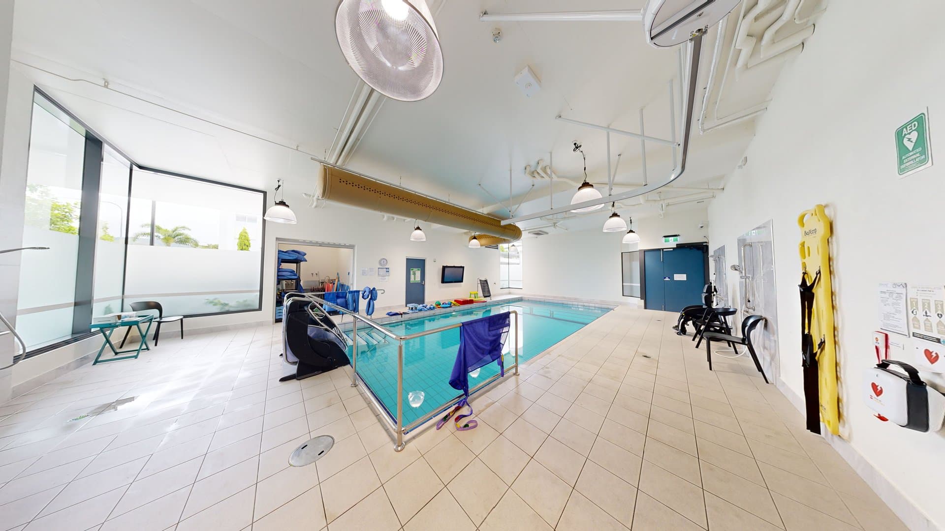 In the middle is a pool equipped with stairs and handrails. To the left is a glass wall, with chairs arranged for seating. To the right is an outdoor shower area. Aquatic wheelchairs are placed against the wall, along with posters and a first aid kit. Swimming accessories are also visible on the other end of the pool. The ceiling and walls are white with an air conditioning vent and stylish light fixtures.