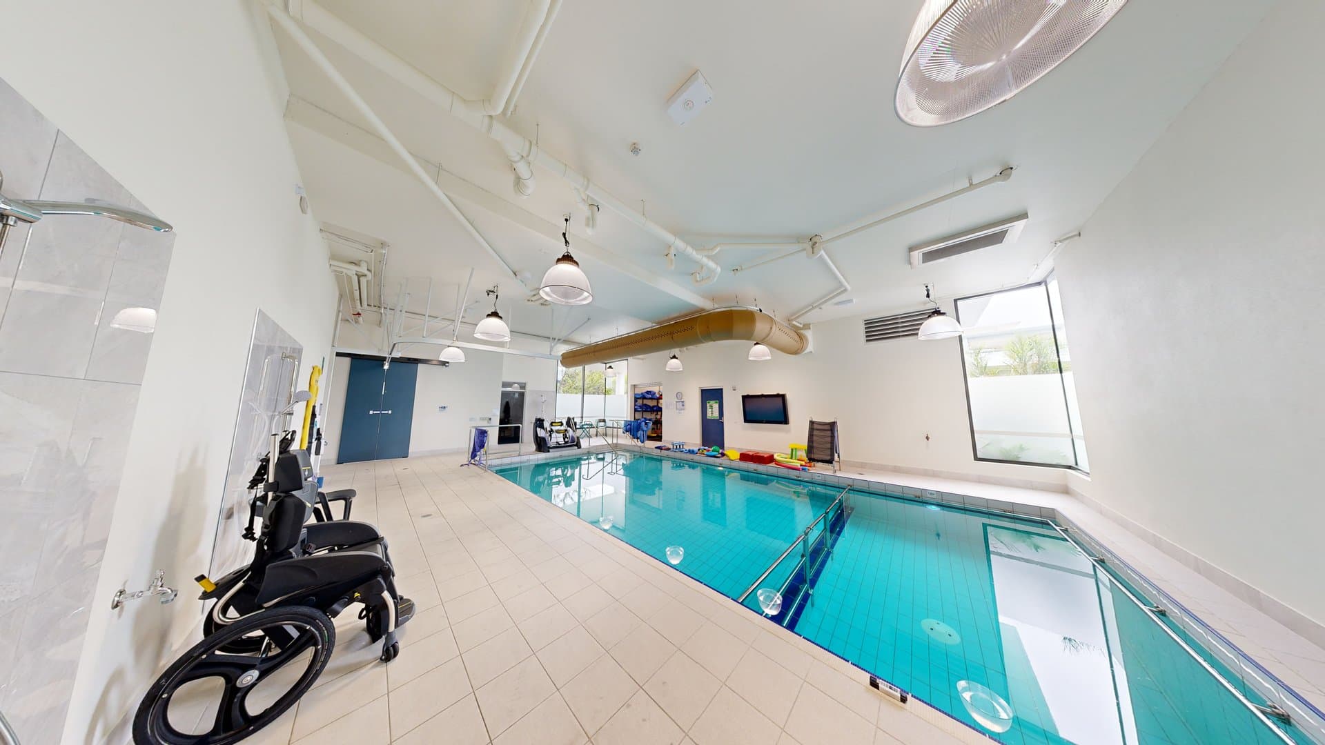 In the centre is a pool with stairs and handrails. On the left is an outdoor shower area. An aquatic wheelchair is placed nearby. Swimming accessories are seen at the opposite end. The ceiling and walls are white with an air conditioning vent and stylish light fixtures. Two glass windows offer an outside view.