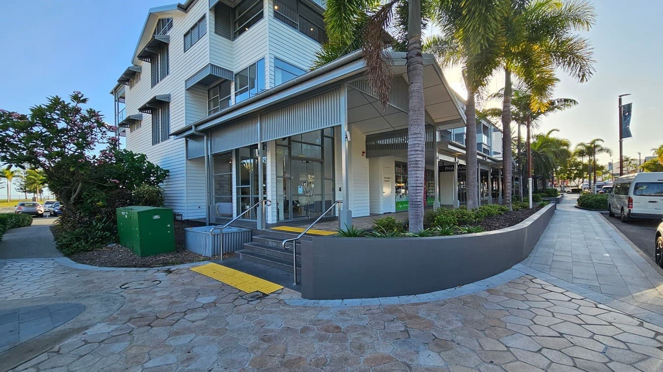 A modern building with a white exterior and large windows is on the left. It has a small set of steps leading to the entrance, with a yellow tactile paving strip. A green utility box and lush plants are nearby. On the right, a row of palm trees lines a paved walkway, with parked vehicles along the street. The pavement is made of hexagonal tiles in various shades. The building has a corrugated metal awning, and the sun is shining through the trees.