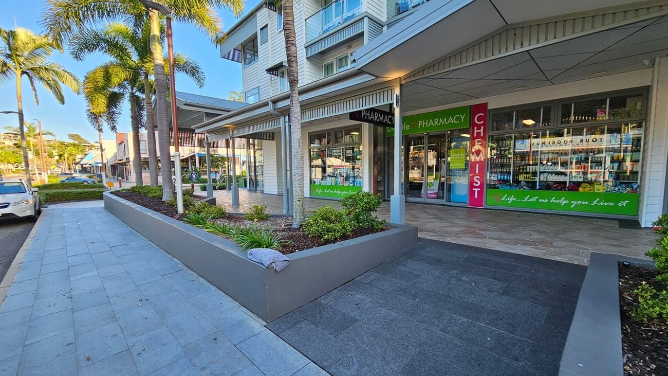 A street scene with a pharmacy on the right, featuring large glass windows displaying products. The entrance is marked by signs reading "PHARMACY" and "CHEMIST" in green and red. In the foreground, a raised garden bed with small plants and a palm tree is visible. On the left, a paved walkway runs alongside parked cars and more palm trees. The building has a modern design with a covered walkway and multiple storeys.