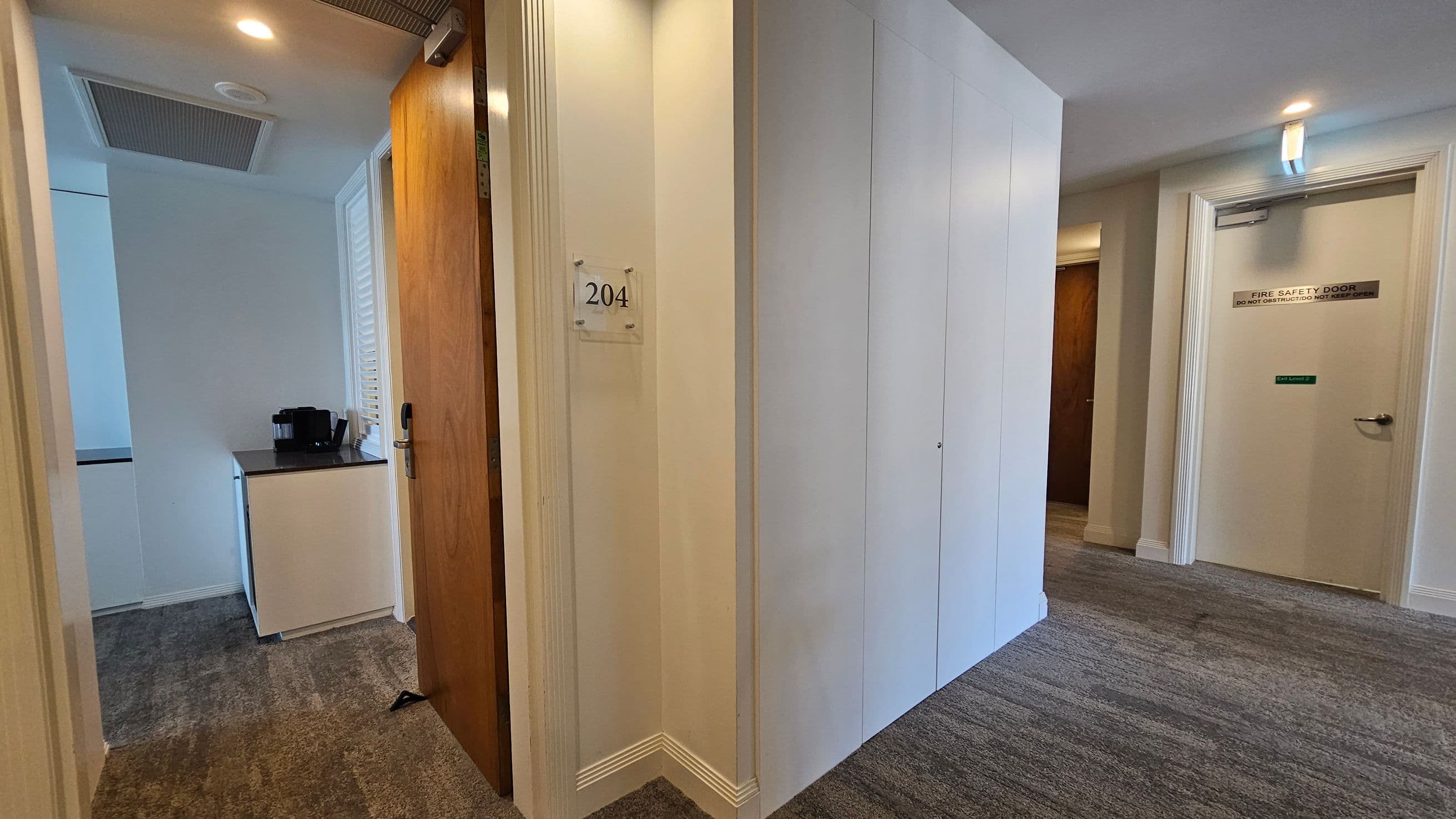 A hallway with a grey and green patterned carpet. On the left, a wooden door is slightly open, revealing a small room with a black coffee machine on a white cabinet. The door has a sign with the number "204". On the right, a white door marked "Fire Safety Door" is closed. The walls are light-coloured, and the ceiling is white with recessed lighting.