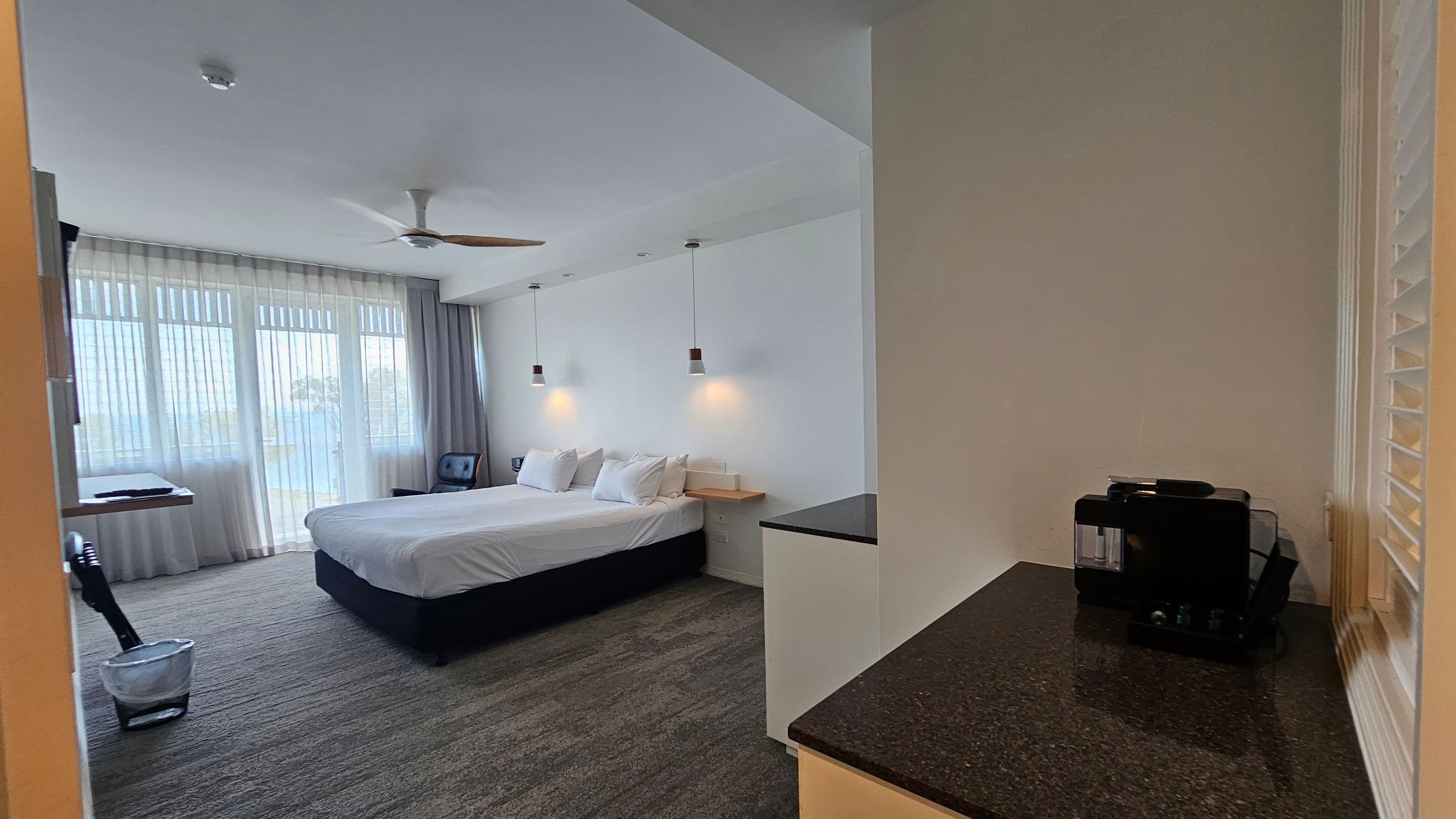 A hotel room with a large bed in the centre, adorned with white bedding and pillows. Two pendant lights hang above the bed. On the left, a large window with sheer curtains allows natural light to enter. A small table and chair are near the window. On the right, a black coffee machine sits on a dark countertop. The floor is carpeted in a grey and green pattern, and a ceiling fan is above the bed.