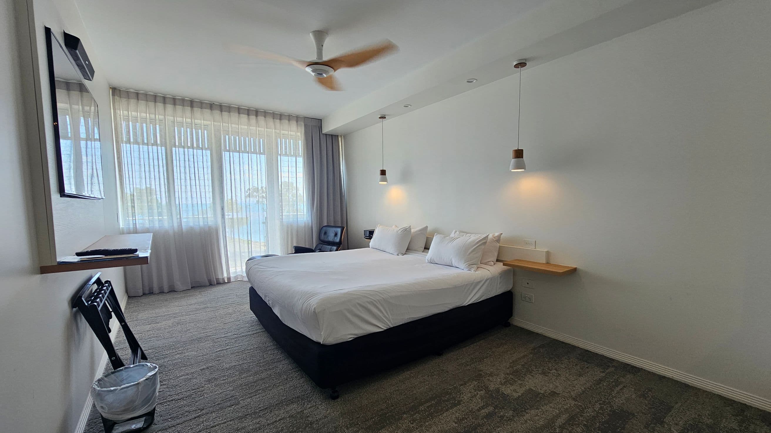 A modern hotel room with a large bed in the centre, adorned with white linens and pillows. Above the bed, two pendant lights hang from the ceiling. On the right, a small wooden shelf is attached to the wall. The left side features a wall-mounted TV above a folding chair and a waste bin. Large windows with sheer curtains allow natural light to fill the room. The floor is carpeted in a mix of grey and green tones, and a ceiling fan is visible above.
