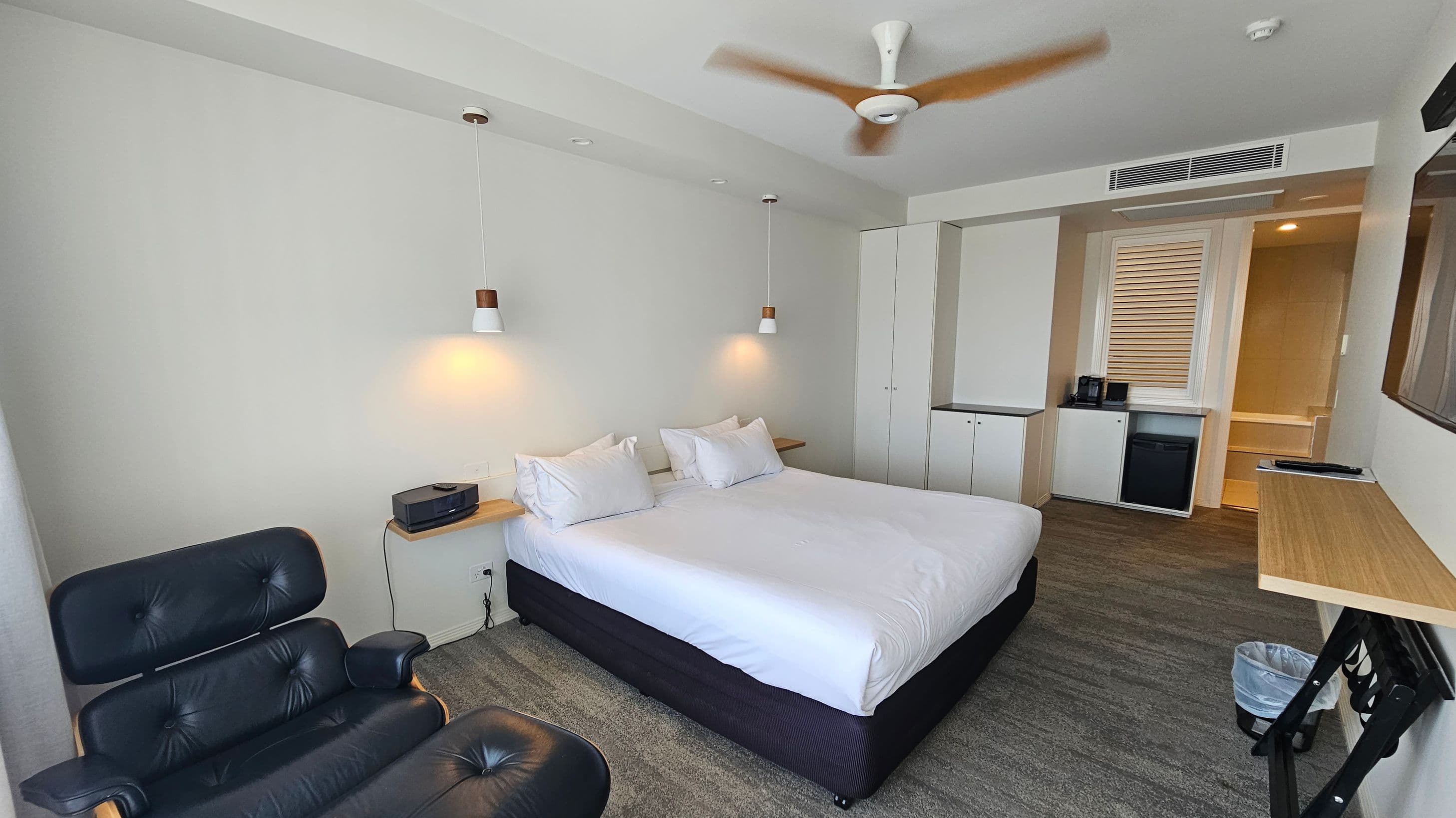 A modern hotel room with a large bed in the centre, flanked by two hanging pendant lights. On the left, there's a black leather chair. To the right, a small kitchenette area with a mini fridge, coffee maker, and a cupboard. A wooden desk is mounted on the wall, with a waste bin underneath. The ceiling features a spinning fan, and the floor is carpeted in a mix of grey and green tones.