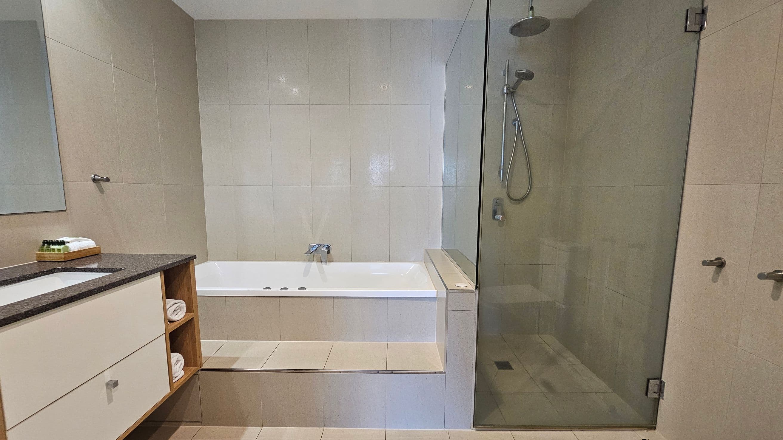 A modern bathroom with a beige tiled floor and walls. On the left, a vanity unit with a dark countertop holds a sink, toiletries, and towels on open shelves. In the centre, a white bathtub is set into a tiled platform. On the right, a glass-enclosed shower area features a rainfall showerhead and a handheld shower. The ceiling is plain white.