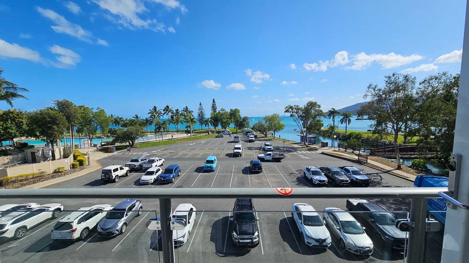 A view from a balcony overlooking a car park with various vehicles, including white and blue cars. Beyond the car park, there are palm trees and a pathway leading to a beach with turquoise water. On the right, a small bridge crosses over a stream surrounded by trees. The scene is bright and sunny, with a clear blue sky.
