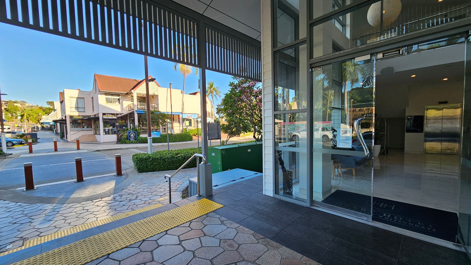 A modern building entrance with glass doors on the right, leading to a lobby with a shiny floor and an elevator. Outside, a paved area with hexagonal tiles and a yellow tactile strip. A small set of stairs with a metal railing descends to the left. Across the street, there are shops with red-tiled roofs and a few parked cars. Greenery, including a bush and a tree, is visible near the entrance.
