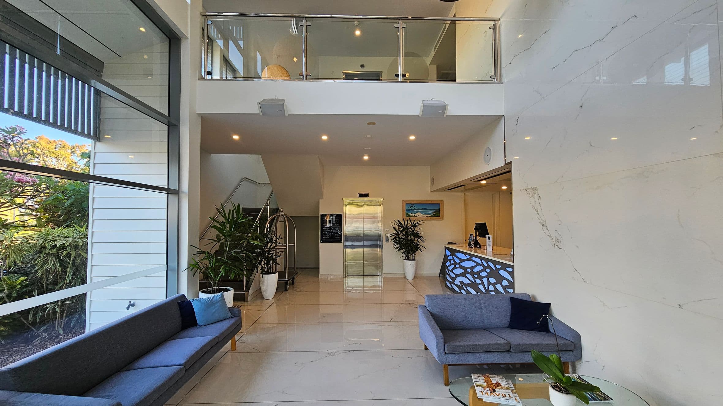 A modern lobby with a high ceiling and a mezzanine level. On the left, large windows reveal lush greenery outside. A long grey sofa with blue cushions is positioned against the window. In the centre, a lift is flanked by potted plants and a staircase. On the right, a reception desk with a decorative panel is accompanied by a grey sofa and a glass coffee table with magazines and a plant. The floor is polished and light-coloured, complementing the sleek, contemporary design.
