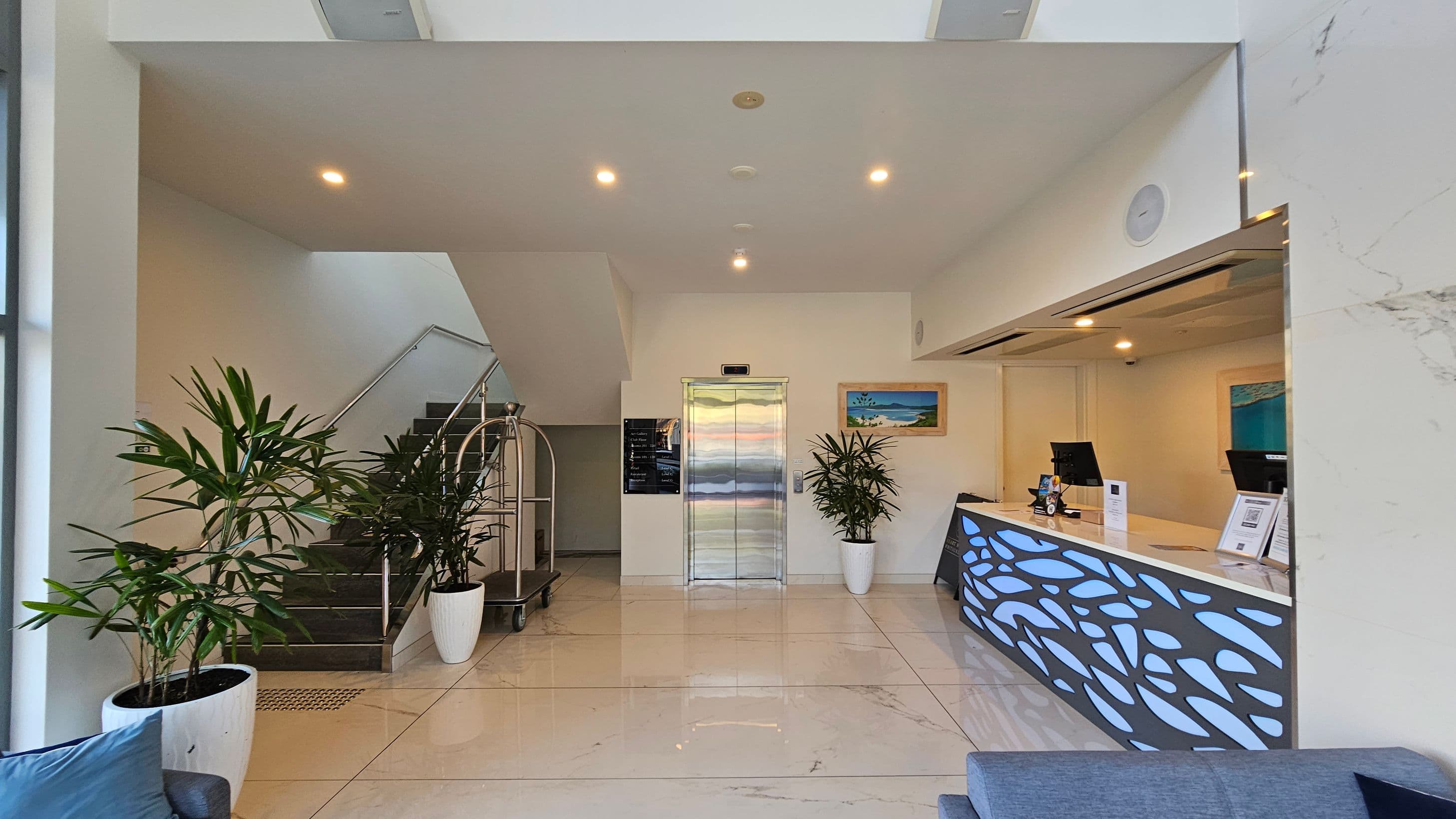 A modern lobby area with a staircase on the left, featuring metal railings and potted plants. In the centre, there's a lift with a metallic door, flanked by more plants and a wall-mounted painting. On the right, a reception desk with a decorative front panel, two computer monitors, and informational materials. The floor is tiled in a light colour, and the ceiling has recessed lighting.