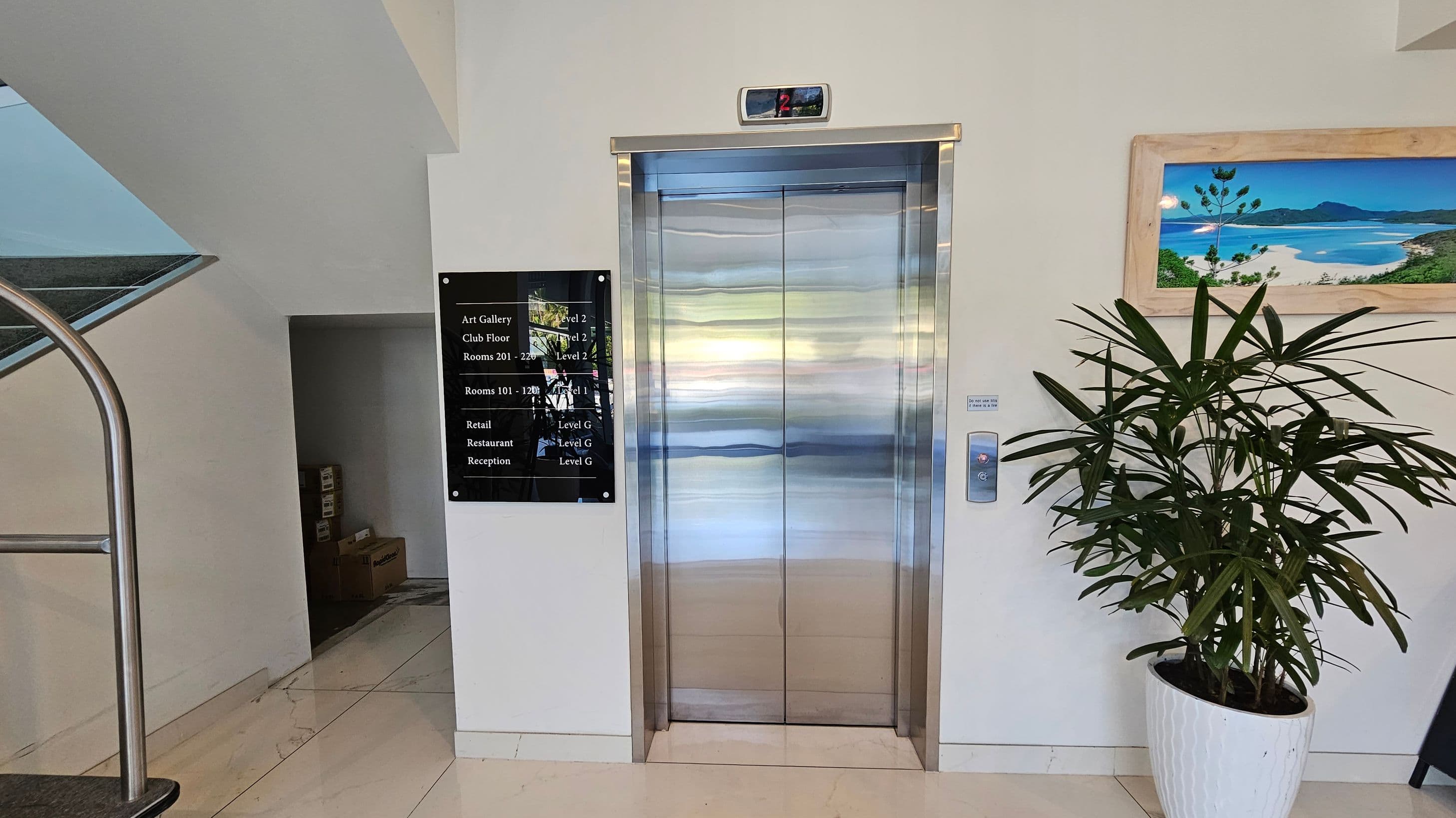 A modern lift with shiny metallic doors is centred on a light-coloured wall. Above the lift, a digital display shows the number "2". To the left, a black directory sign lists various locations and levels. A staircase with a metal railing is partially visible. On the right, a potted plant with long, slender leaves stands next to a framed landscape picture. The floor is tiled in a light colour.