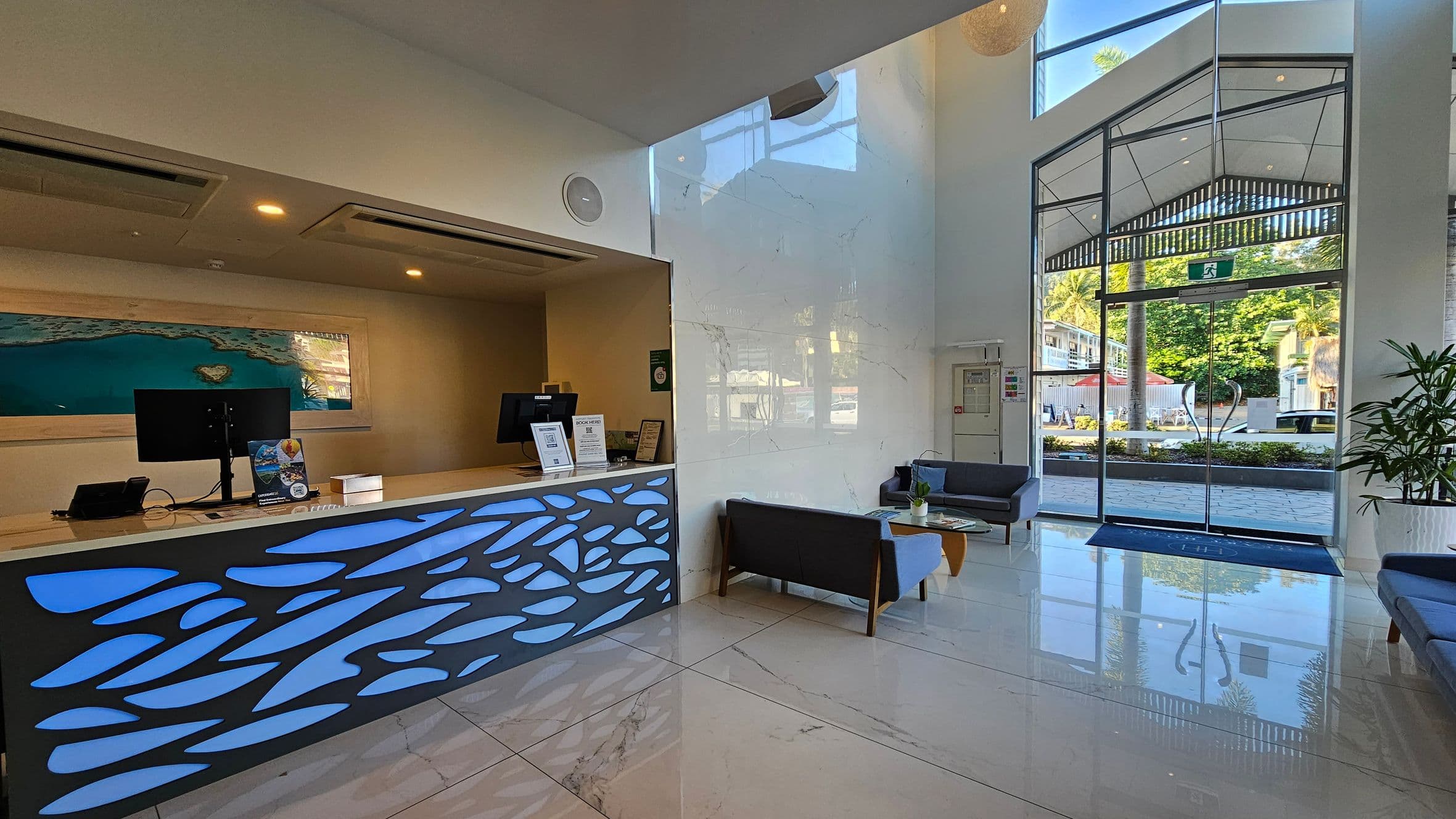 A modern reception area with a sleek desk on the left, featuring a blue abstract design and two computer monitors. Behind the desk is a large framed image of a coastal scene. On the right, there are glass doors leading outside, with a view of greenery and buildings. Two grey sofas and a wooden coffee table are positioned near the entrance. The floor is polished marble, and the ceiling is high with recessed lighting.
