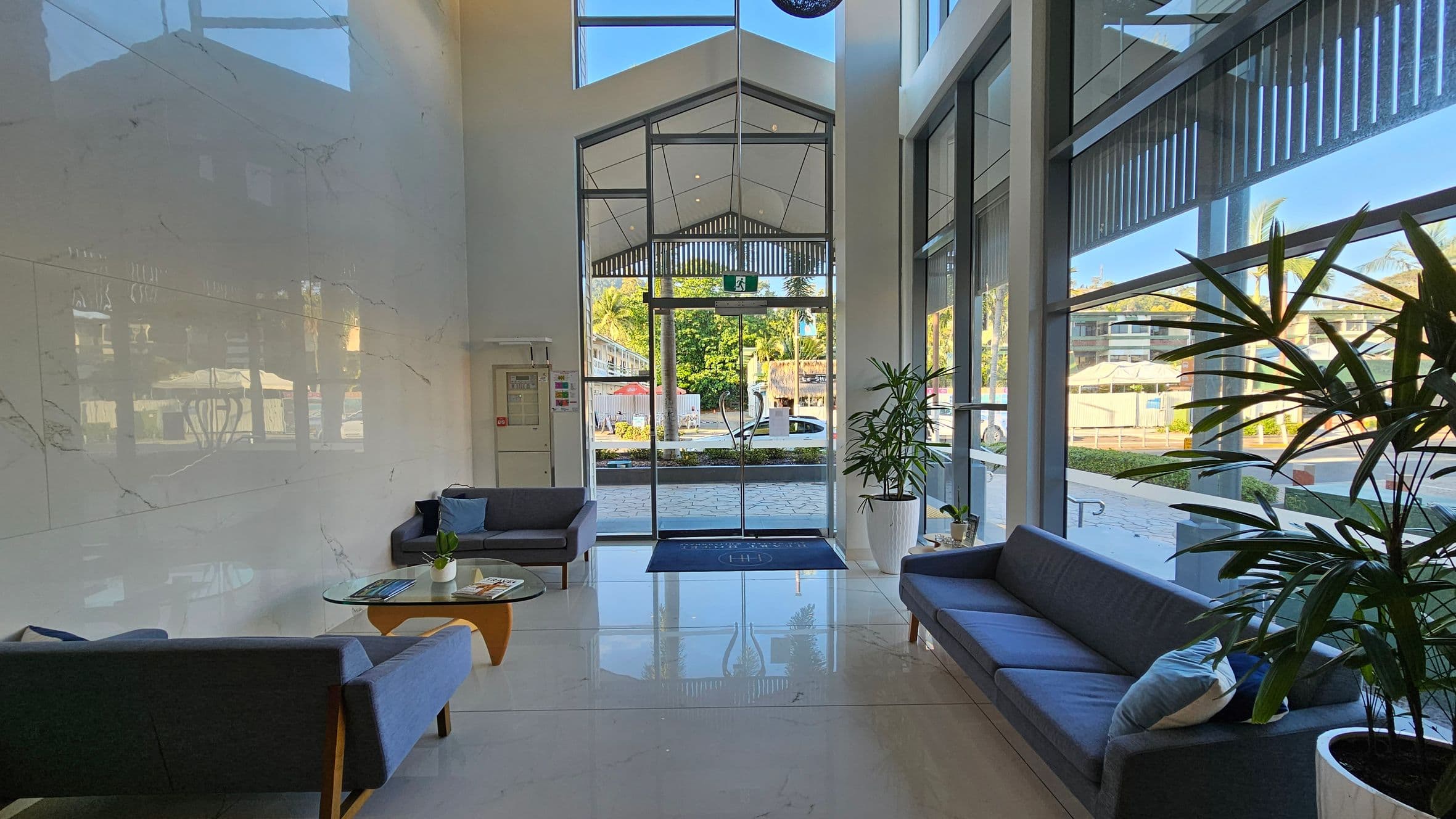 A modern lobby with a high ceiling and large glass entrance doors. On the left, there are two grey sofas with blue cushions and a glass coffee table with magazines and a small plant. On the right, another grey sofa with a cushion is next to a large potted plant. The floor is shiny and tiled, reflecting the furniture and natural light. Outside, greenery and a street are visible through the glass.