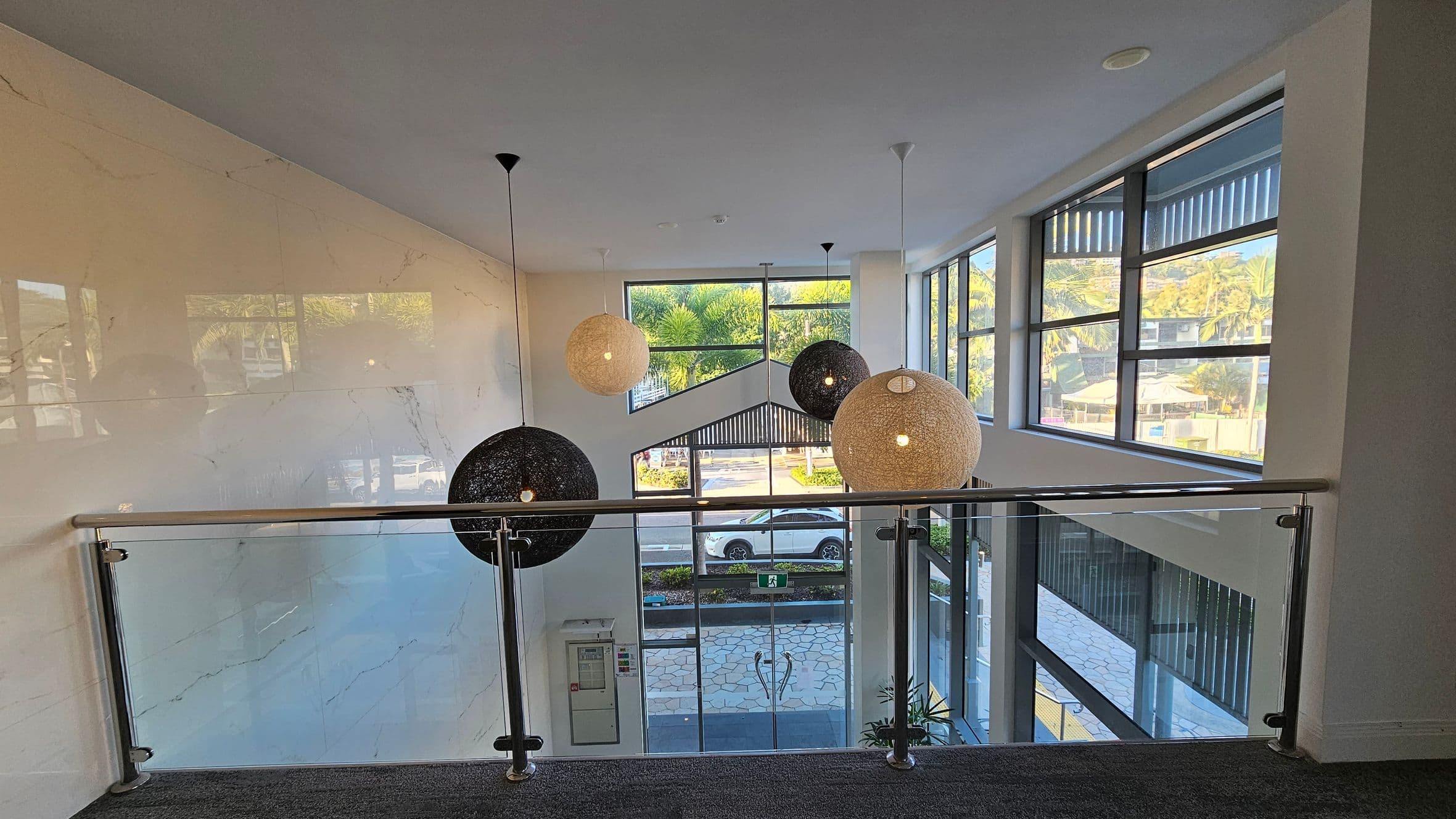 A modern interior with large windows on the right, allowing natural light to illuminate the space. In the centre, three spherical pendant lights hang from the ceiling, two black and one beige. A glass railing with metal supports runs across the foreground. The floor is carpeted in a dark grey tone. Outside, a view of a street with a parked car and greenery is visible through the windows.