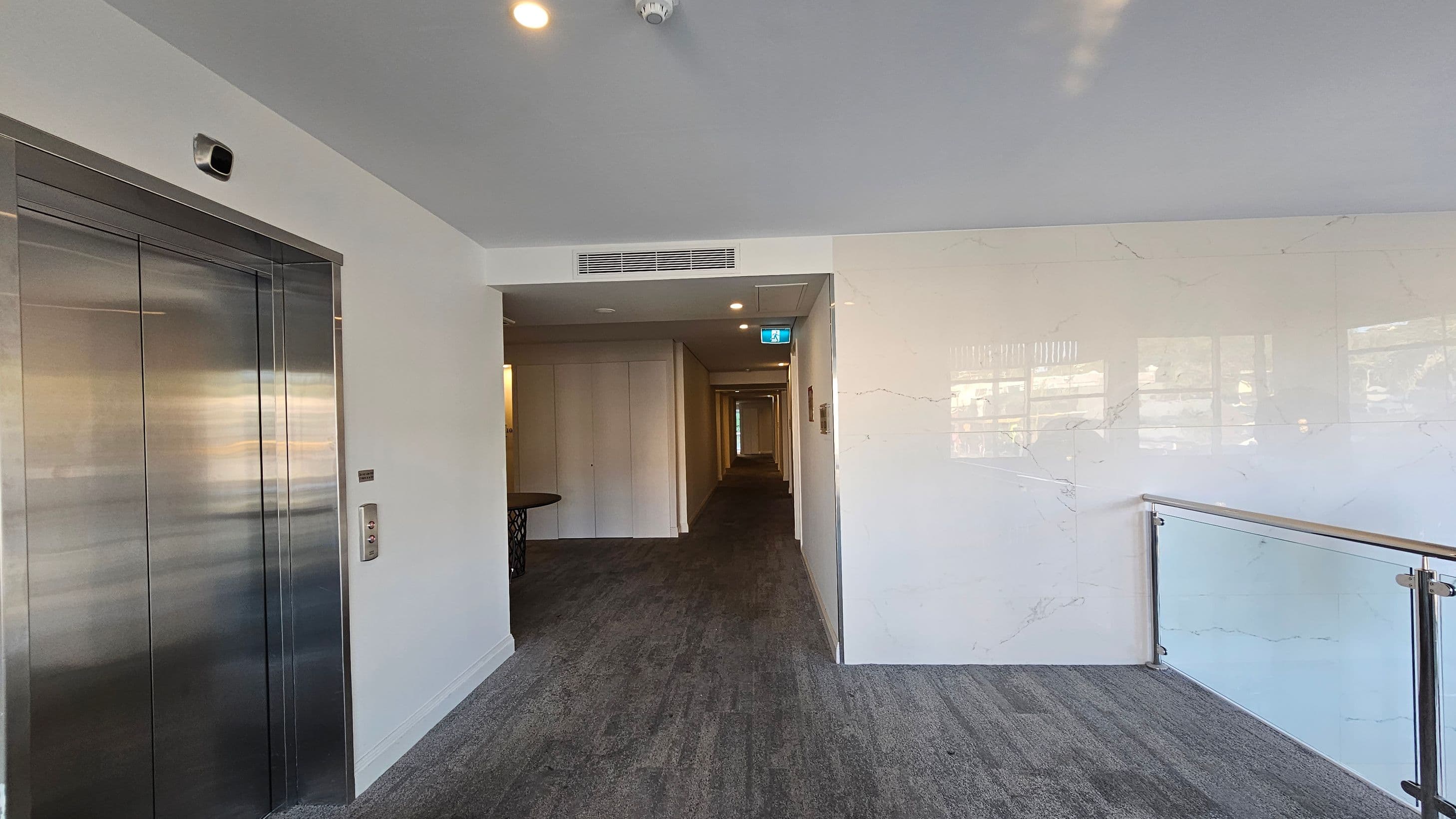 On the left, there is a metallic lift door with a control panel beside it. In the centre, a hallway extends into the distance, with a table visible near the entrance. The right side features a glass railing with a wooden handrail, overlooking a lower level. The floor is carpeted in a grey tone, and the ceiling is white with recessed lighting and a smoke detector.