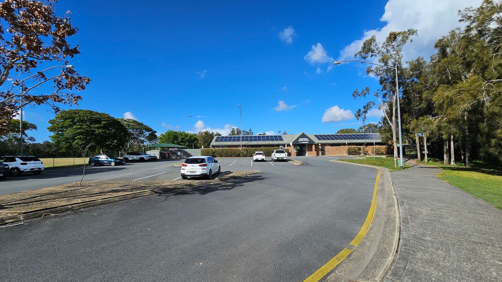 A car park with several vehicles is in the foreground, with a paved road curving to the right. In the centre, a brick building with solar panels on the roof is visible. To the left, a grassy area with trees lines the road, while more trees are on the right. A lamppost stands near the pavement on the right side. The sky is clear with a few clouds.