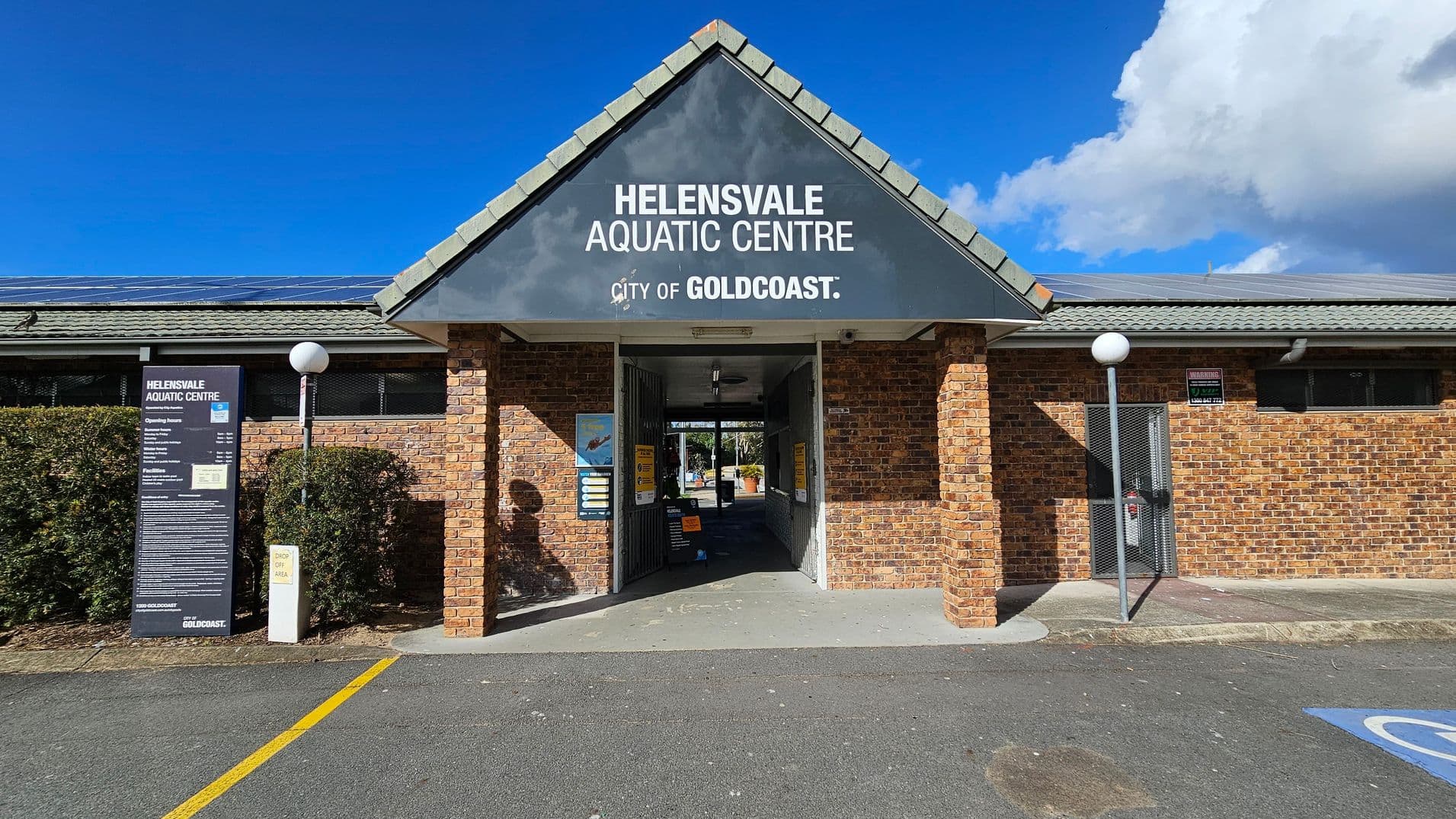 A brick building with a triangular roof features a sign reading "Helensvale Aquatic Centre, City of Gold Coast." The entrance is in the centre, with open gates leading inside. On the left, a tall sign displays information about the centre, surrounded by bushes. Two spherical lamps on poles are positioned on either side of the entrance. The ground is paved with a marked parking area, and the roof has solar panels.
