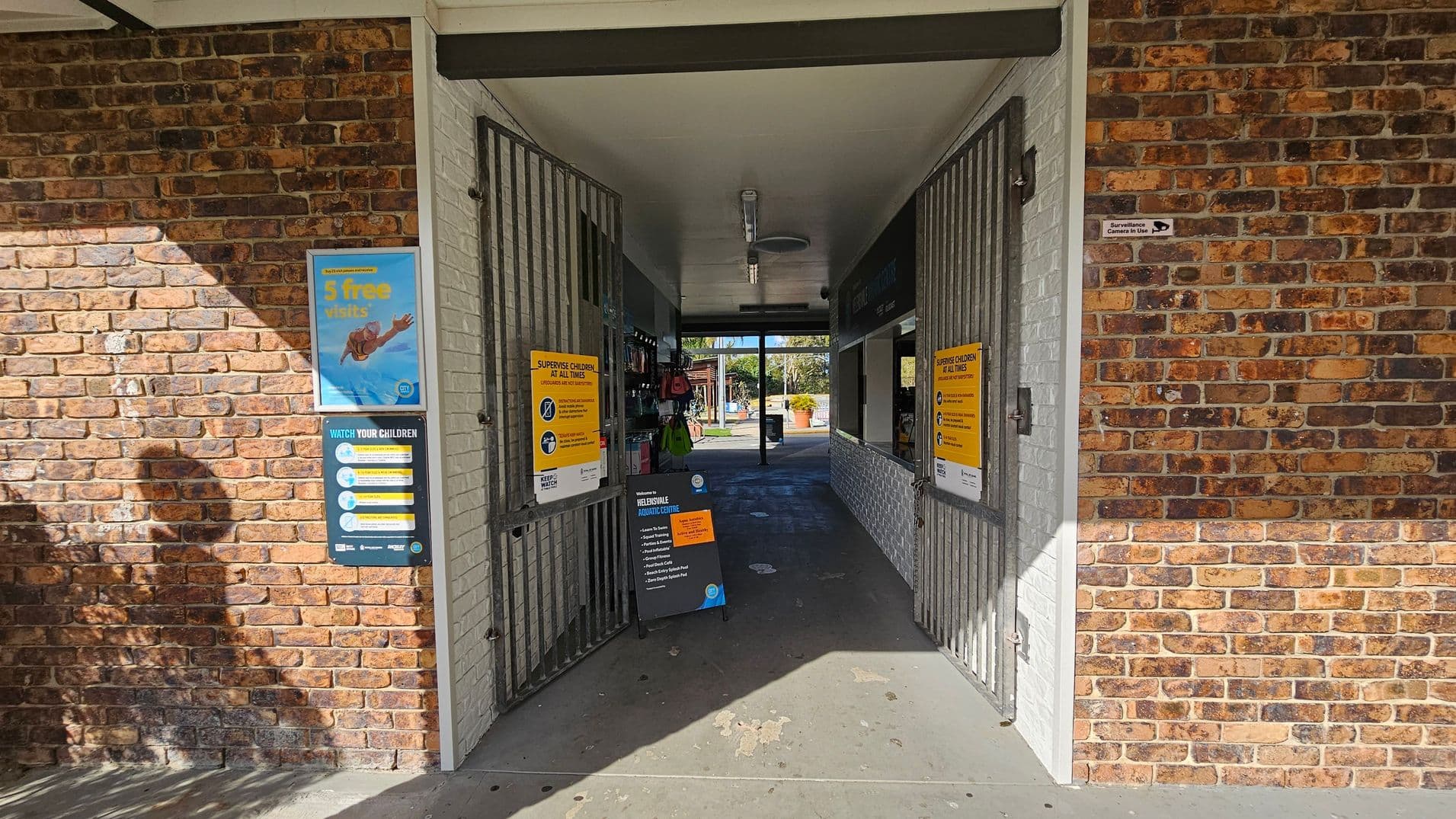 A brick building entrance with two open metal gates. On the left, a poster advertises "5 free visits" and another sign advises to watch children. On the right, a similar sign about supervising children is visible. Inside, a corridor leads to an outdoor area with greenery. The floor is concrete, and the ceiling is white with a light fixture. Shadows of trees are cast on the left wall.