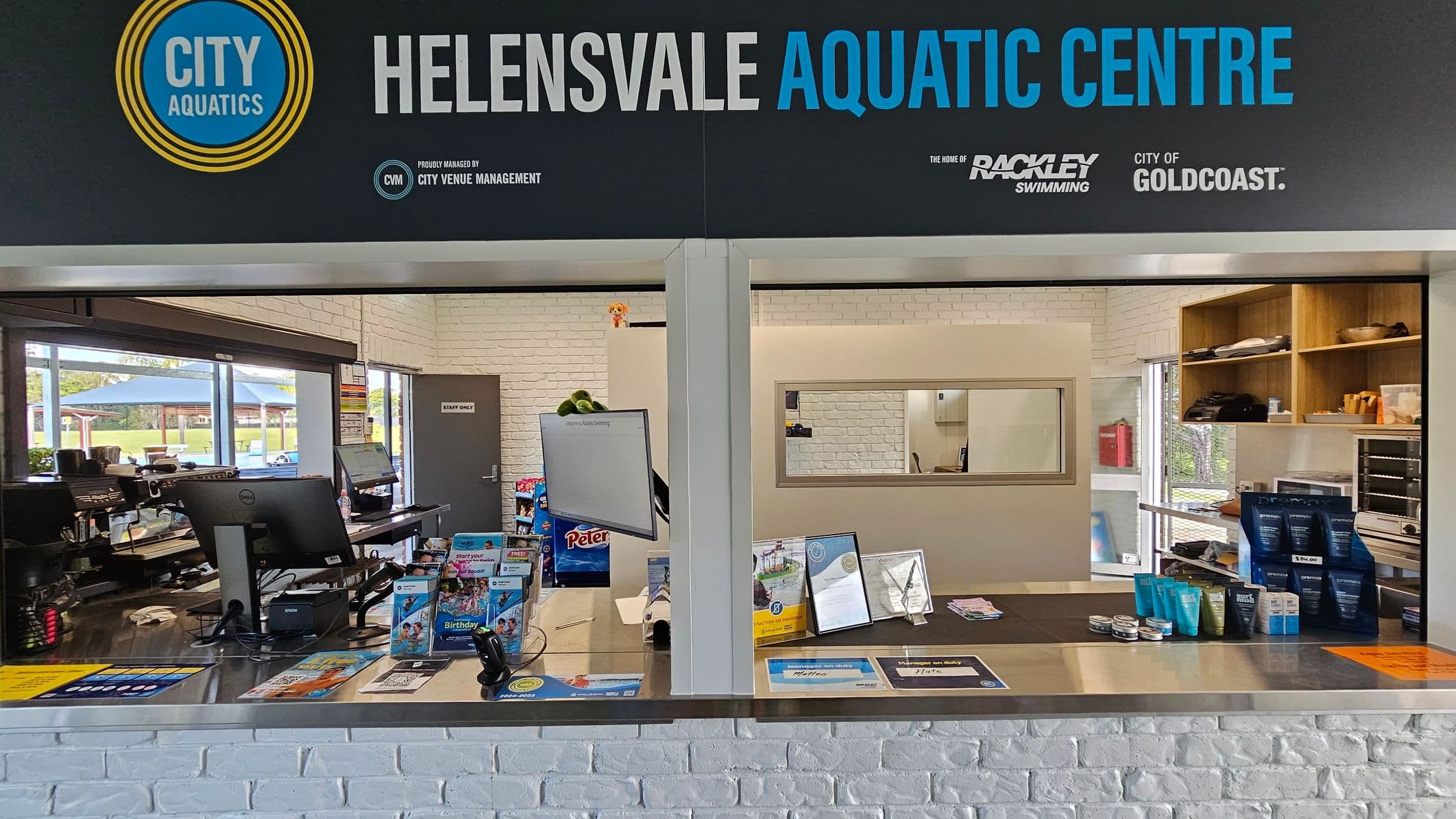 Reception area of the Helensvale Aquatic Centre. The foreground features a counter with brochures, a computer monitor, and various products on display. The background shows a white brick wall with a "Staff Only" door and shelves containing items. To the left, there is a window with a view of an outdoor area with tables and umbrellas. The ceiling is not visible.
