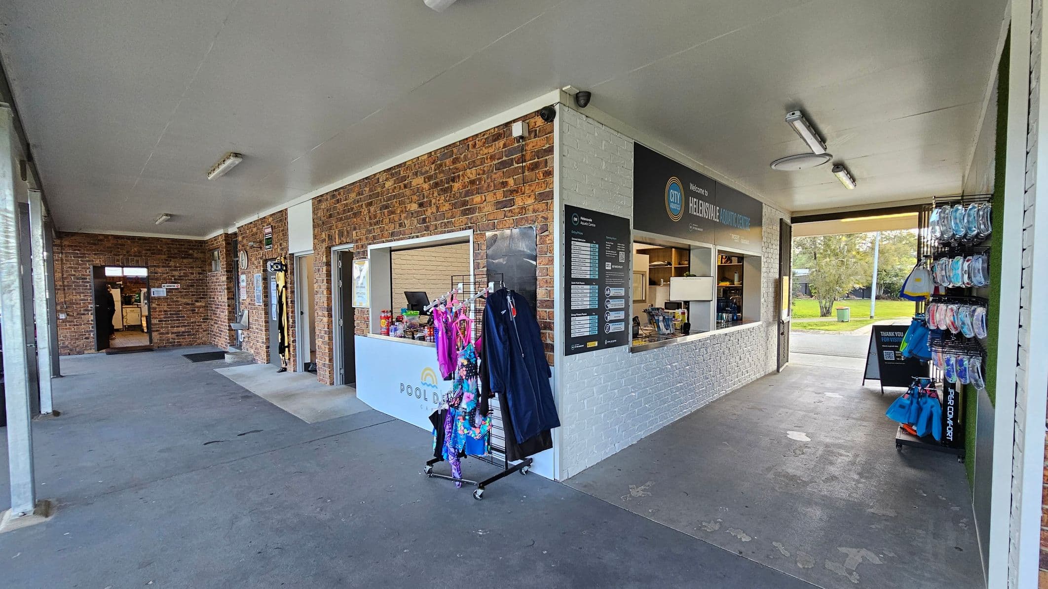 A corridor with a concrete floor and a white ceiling. On the left, a brick wall with an open doorway leads to another room. In the middle, a rack displays colourful swimwear, and a counter with a sign reads "Helensvale Aquatic Centre." On the right, shelves hold swimming accessories like goggles and fins. The background shows a grassy area with trees visible through an open space. Fluorescent lights are mounted on the ceiling.