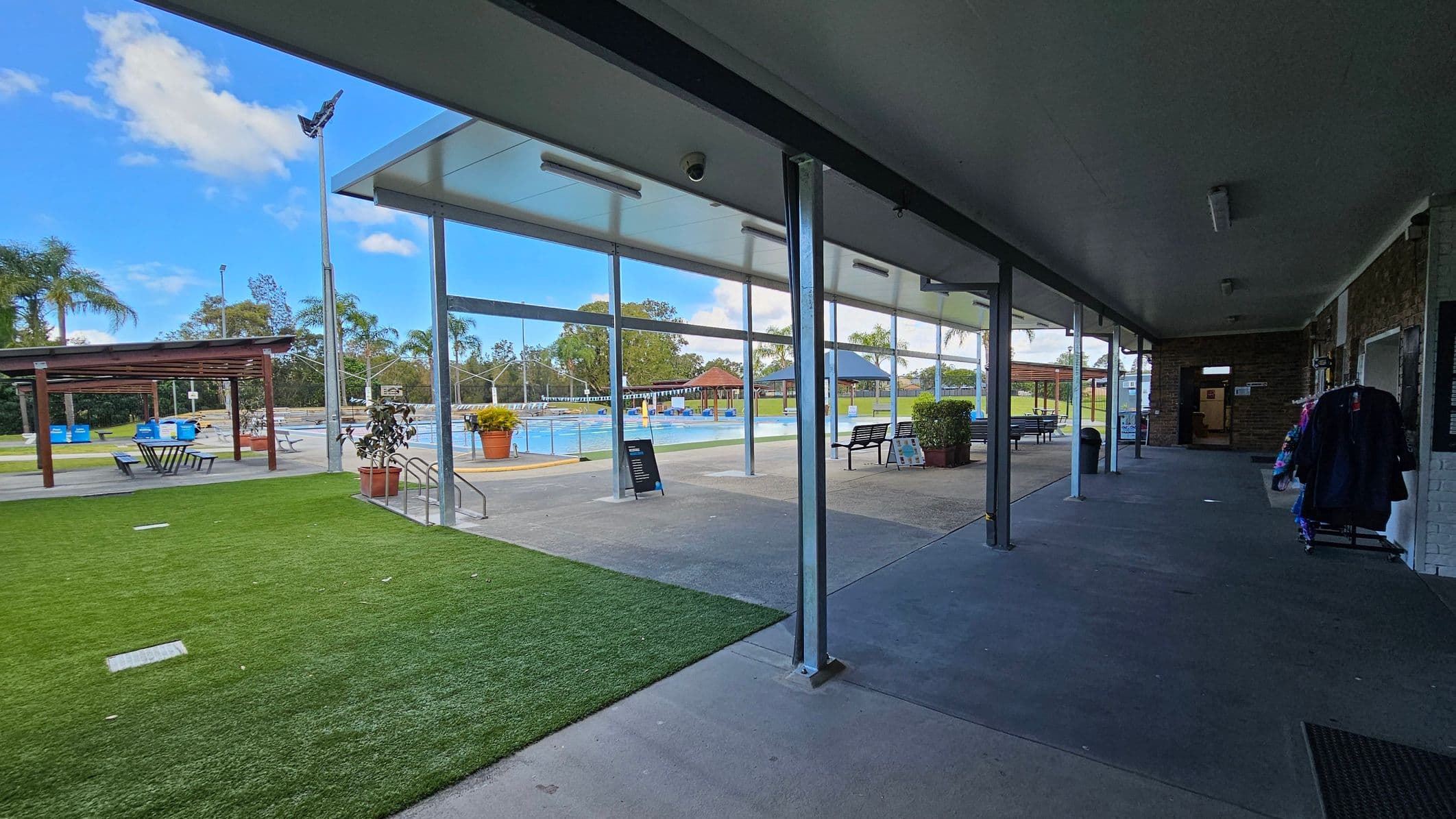 A covered walkway with a concrete floor on the right, leading to a building entrance. On the left, there's a grassy area with a wooden pergola and picnic tables. Beyond the walkway, a swimming pool is visible, surrounded by potted plants and benches. The area is open and spacious, with trees and a clear sky in the background. The ceiling of the walkway is white with lighting fixtures.