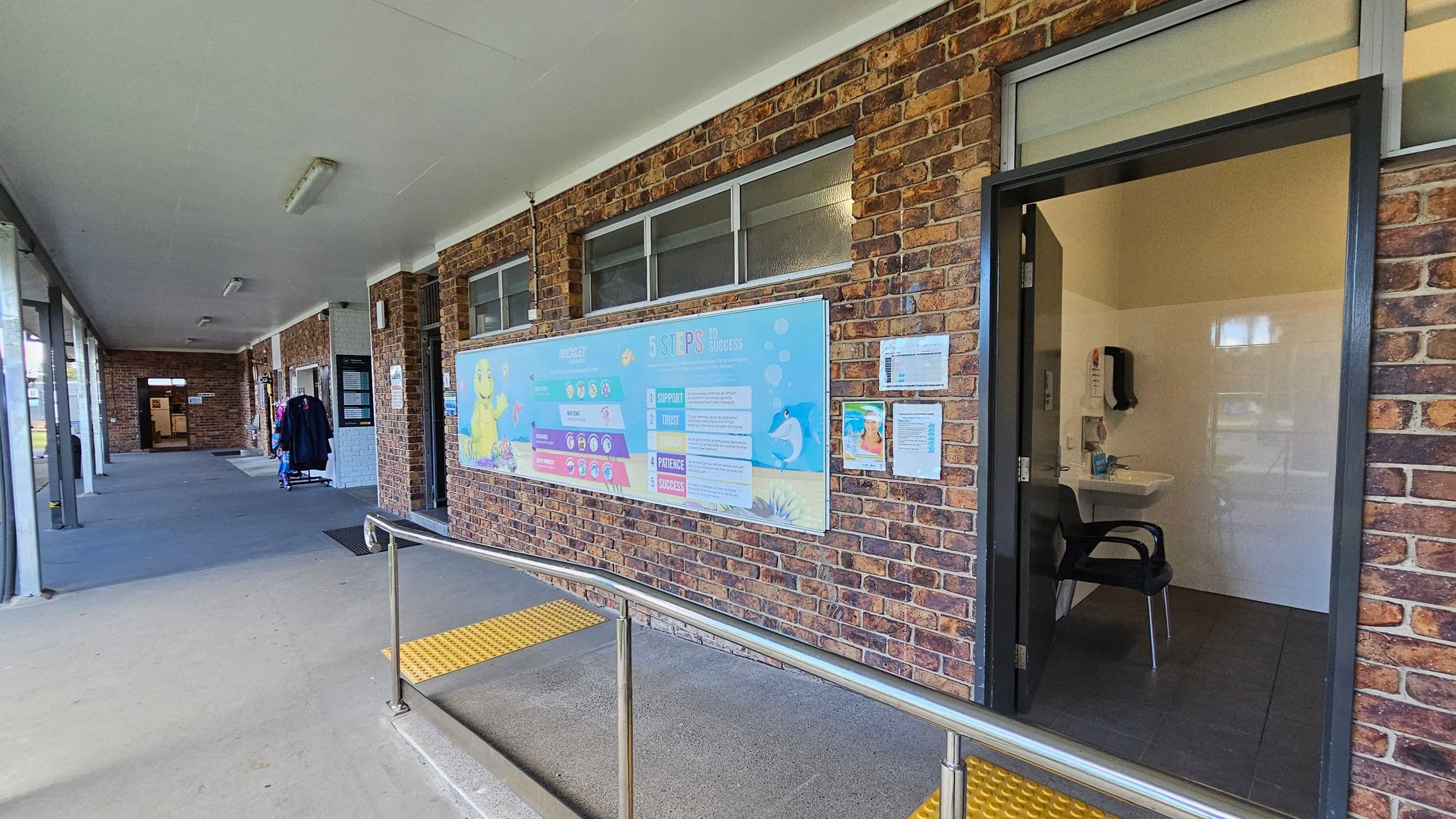 A corridor with a brick wall on the right featuring a colourful poster titled "5 Steps to Success". Below the poster, a door is open, revealing a small room with a chair and a sink. On the left, the corridor extends with a few coats hanging on a rack. The floor is concrete with a yellow tactile strip near the door, and a metal handrail runs along the edge. The ceiling is white with fluorescent lights.