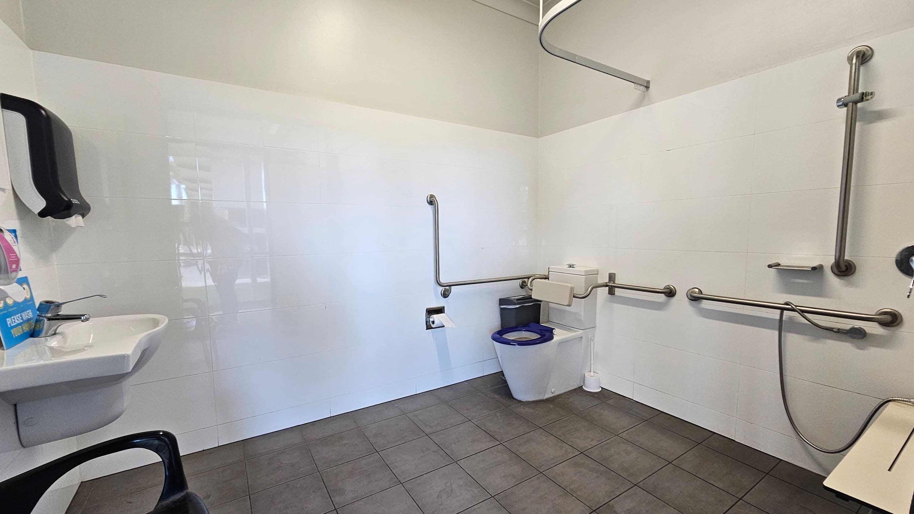Accessible bathroom with white tiled walls and grey tiled floor. On the left, a wall-mounted sink with a soap dispenser above it. In the centre, a toilet with a blue seat, surrounded by metal grab bars. A toilet paper holder is mounted on the wall. On the right, a shower area with additional grab bars and a handheld showerhead. A foldable shower seat is visible. The ceiling is plain and light-coloured.