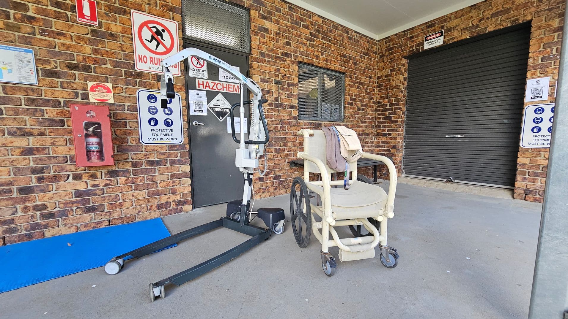 A brick wall with various safety signs, including "No Running" and "Hazchem," is in the background. A fire extinguisher is mounted on the wall. In the foreground, a white mobility chair with large wheels is next to a mechanical lift. A black roller door is on the right, and a blue ramp is on the ground. The ceiling is plain and light-coloured.