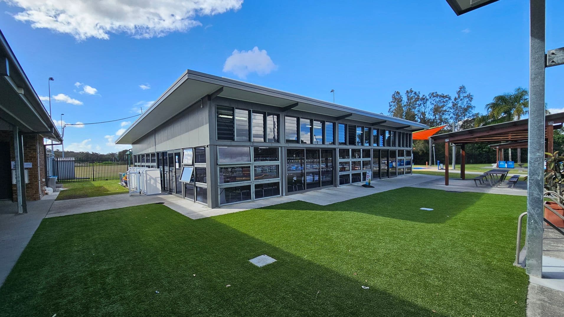 A modern building with large windows on the right, featuring a sloped roof. In front, there's a well-maintained artificial grass area. To the left, a pathway leads to a fenced field. On the right, a wooden pergola with seating is visible. In the background, there are trees and a bright orange shade sail. The ground is a mix of concrete and artificial grass.