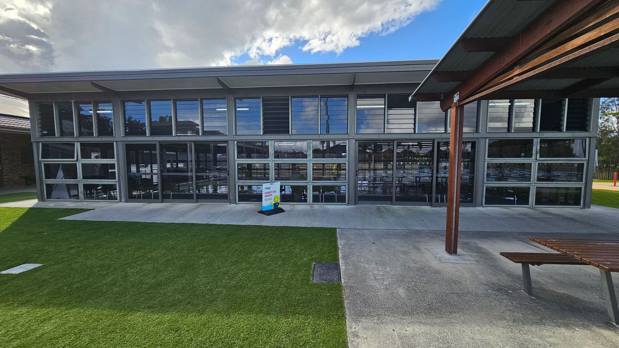 A modern building with large glass windows and louvres spans the centre of the image. In front, a concrete path separates a patch of green grass from the building. A signboard near the entrance reads "Learn to Swim." On the right, a wooden bench and a wooden post supporting a roof are visible. The background shows reflections of trees and structures in the windows.