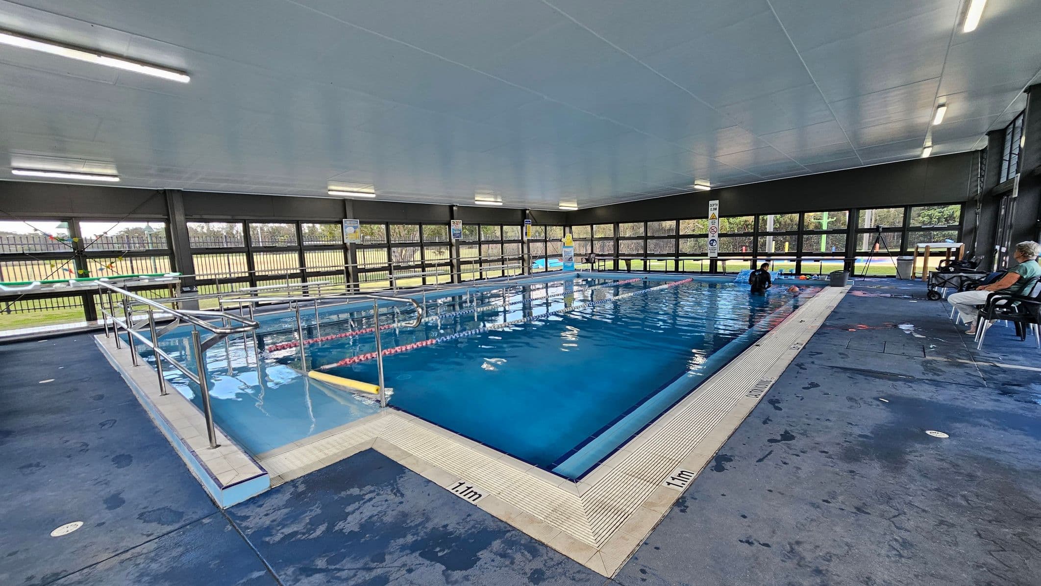 Indoor swimming pool with clear blue water, surrounded by a tiled edge marked with "1.1m" depth indicators. On the left, a metal handrail leads into the pool. The right side has chairs with people seated, observing the pool. Large windows line the background, allowing natural light and views of the outdoor area with grass and trees. The ceiling is white with fluorescent lights, and the floor is a dark, wet surface.